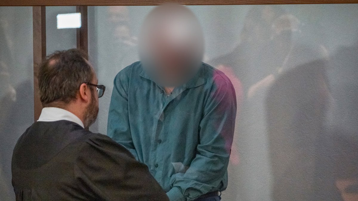 Der Angeklagte steht hinter einer Glaswand im Gerichtssaal. Vor der Wand steht sein Anwalt und sieht ihm ins Gesicht. 