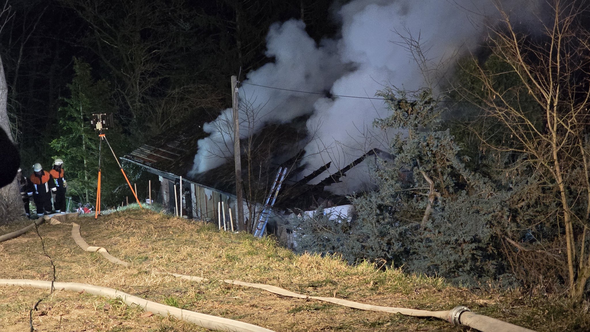 Nach einem Wohnhausbrand in Brennberg im Kreis Regensburg haben Rettungskräfte eine Leiche geborgen. Aus dem beschädigten Dach dringt dichter Rauch. Feuerwehrleute legen Schläuche.