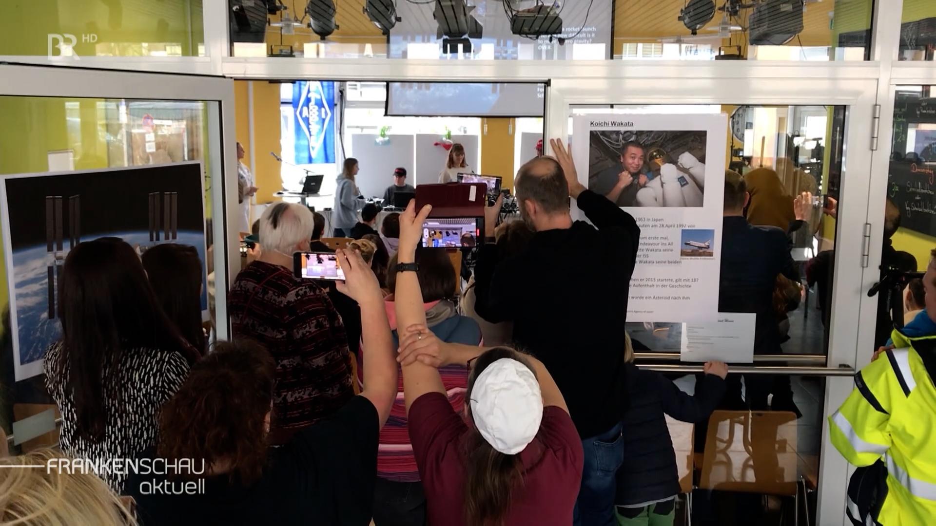 Mehrere Personen halten ihrer Smartphones in die Höhe und fotografieren, im Hintergrund das Foto eines Astronauten. 