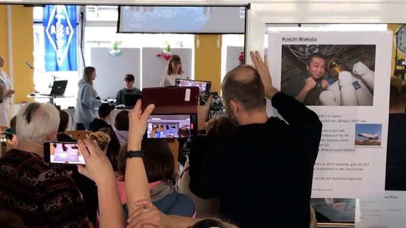 Mehrere Personen halten ihrer Smartphones in die Höhe und fotografieren, im Hintergrund das Foto eines Astronauten. | Bild: BR Mehrere Personen halten ihrer Smartphones in die Höhe und fotografieren, im Hintergrund das Foto eines Astronauten.