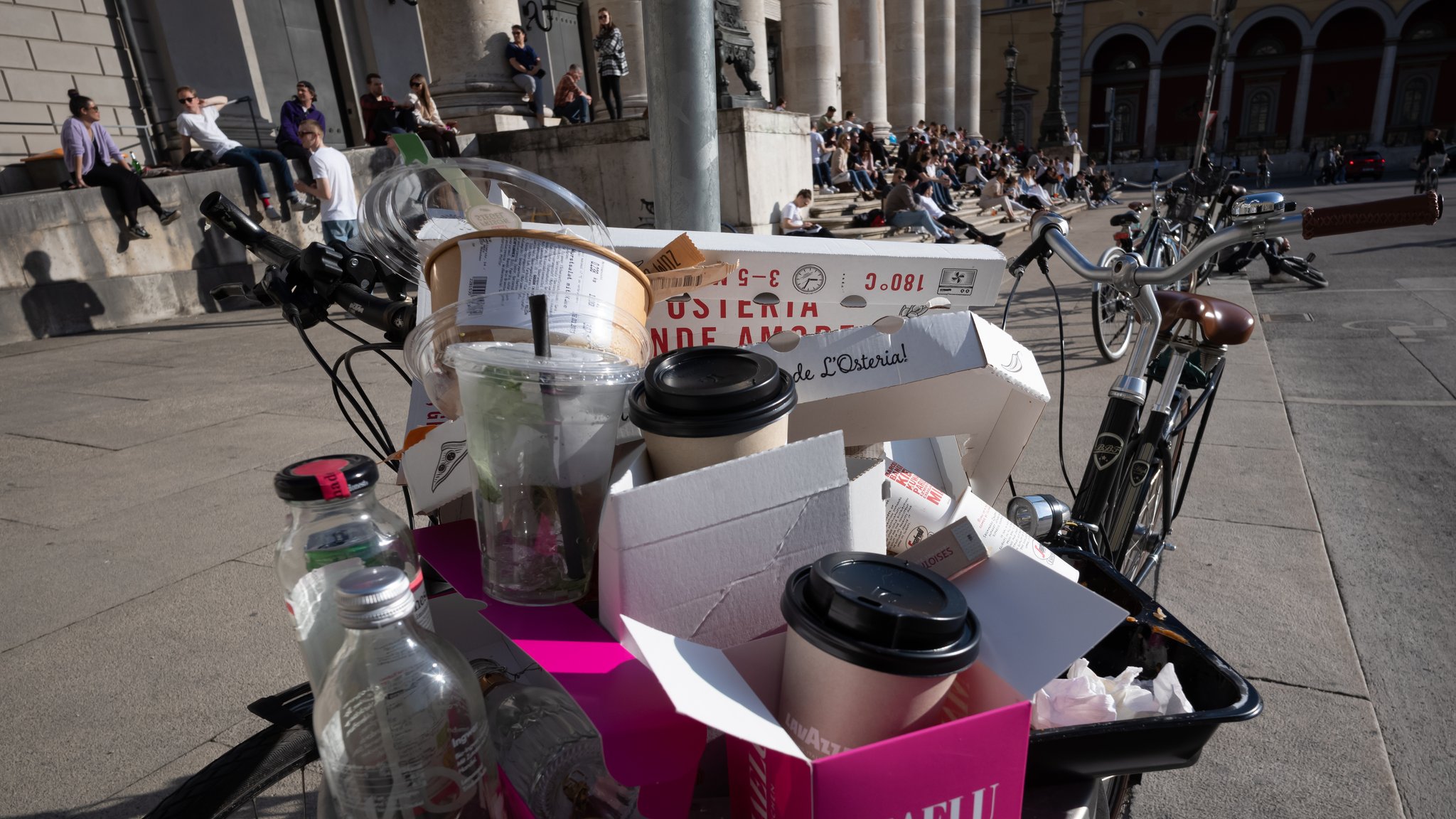 Volle Mülleimer quellen über, während Passanten auf der Freitreppe der bayerischen Staatsoper am Max-Joseph-Platz sitzen.