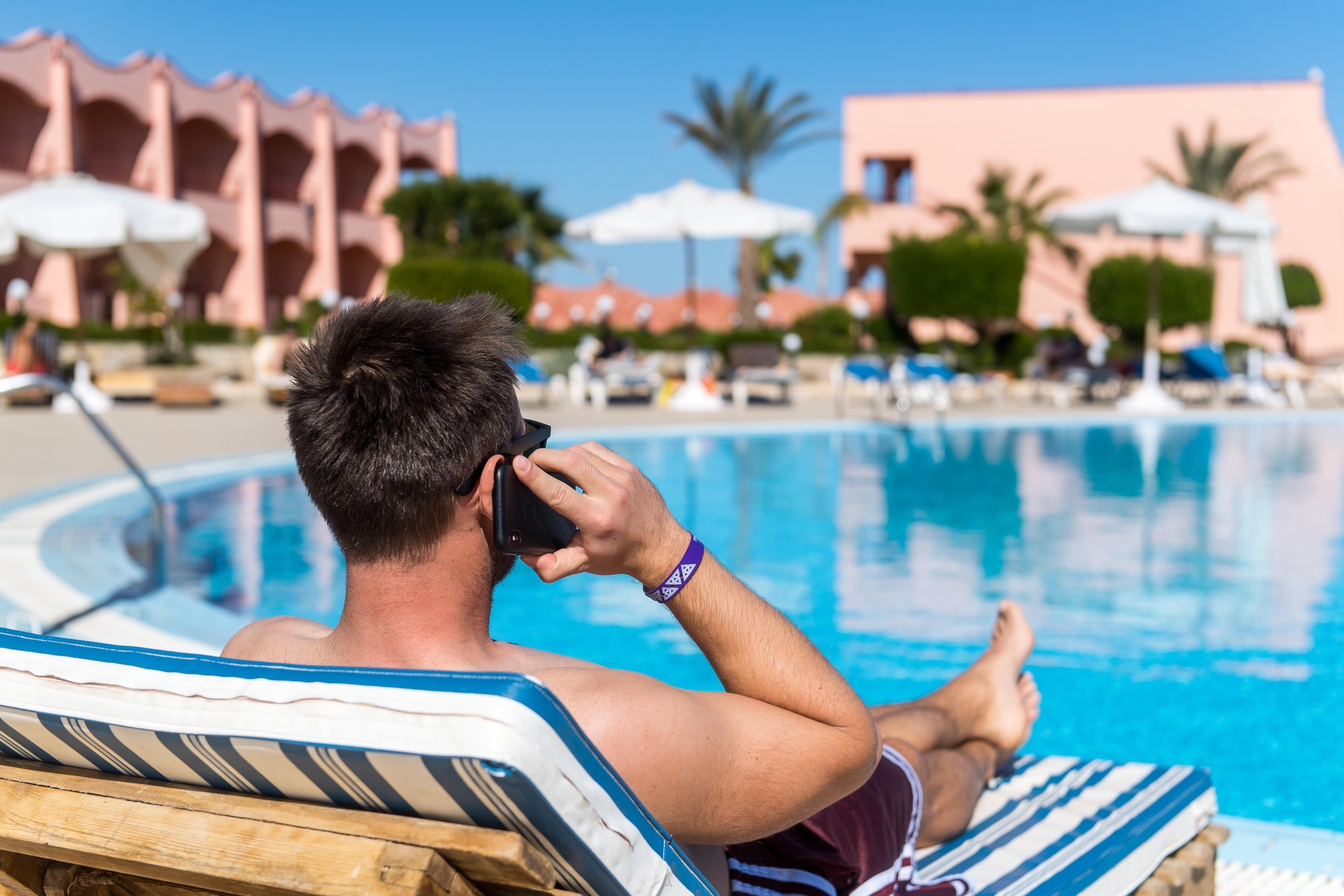 Ein Mann liegt an einem Hotelpool auf einer Sonnenliege und telefoniert (gestellte Szene). 