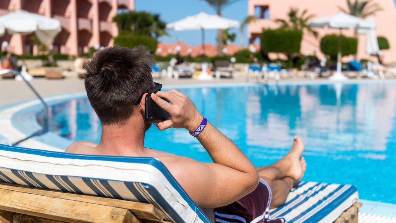 Ein Mann liegt an einem Hotelpool auf einer Sonnenliege und telefoniert (gestellte Szene). 
