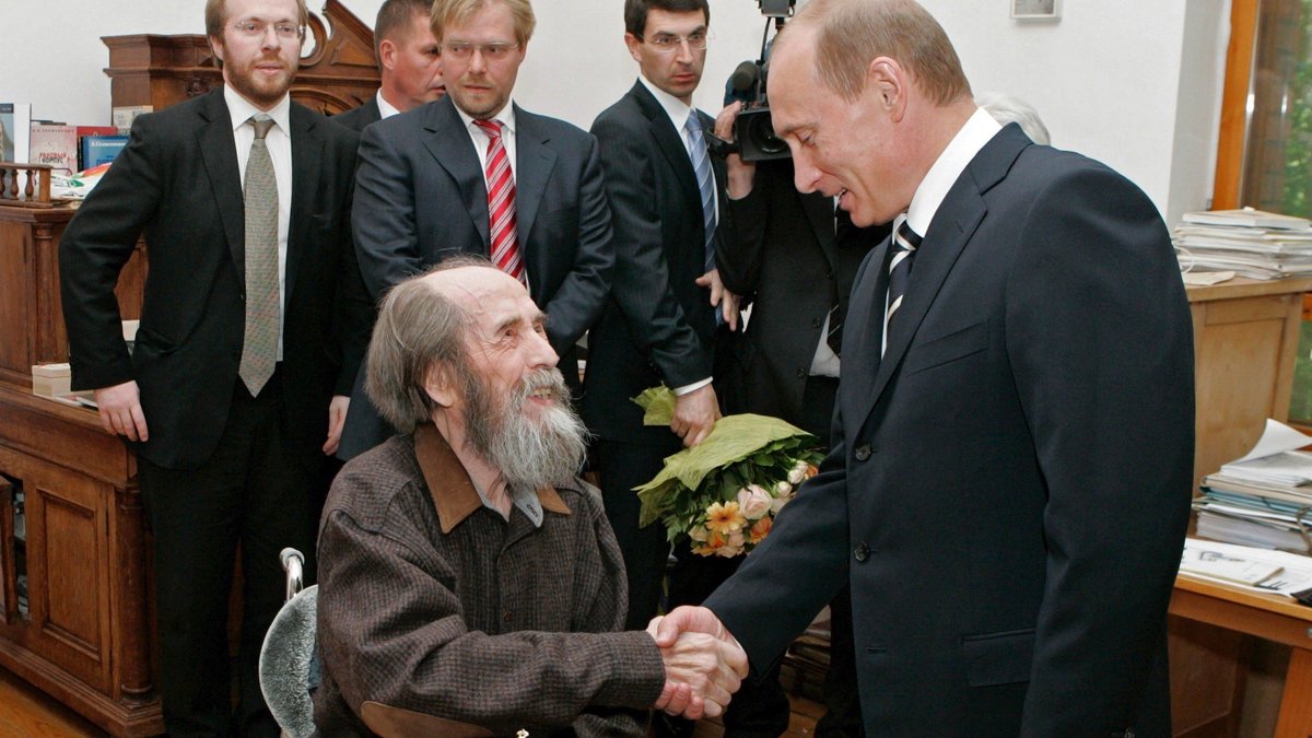 Greiser, bärtiger Schriftsteller sitzt lächelnd an einem Tisch, rechts steht Präsident Putin, der ihm grinsend die Hand schüttelt. 