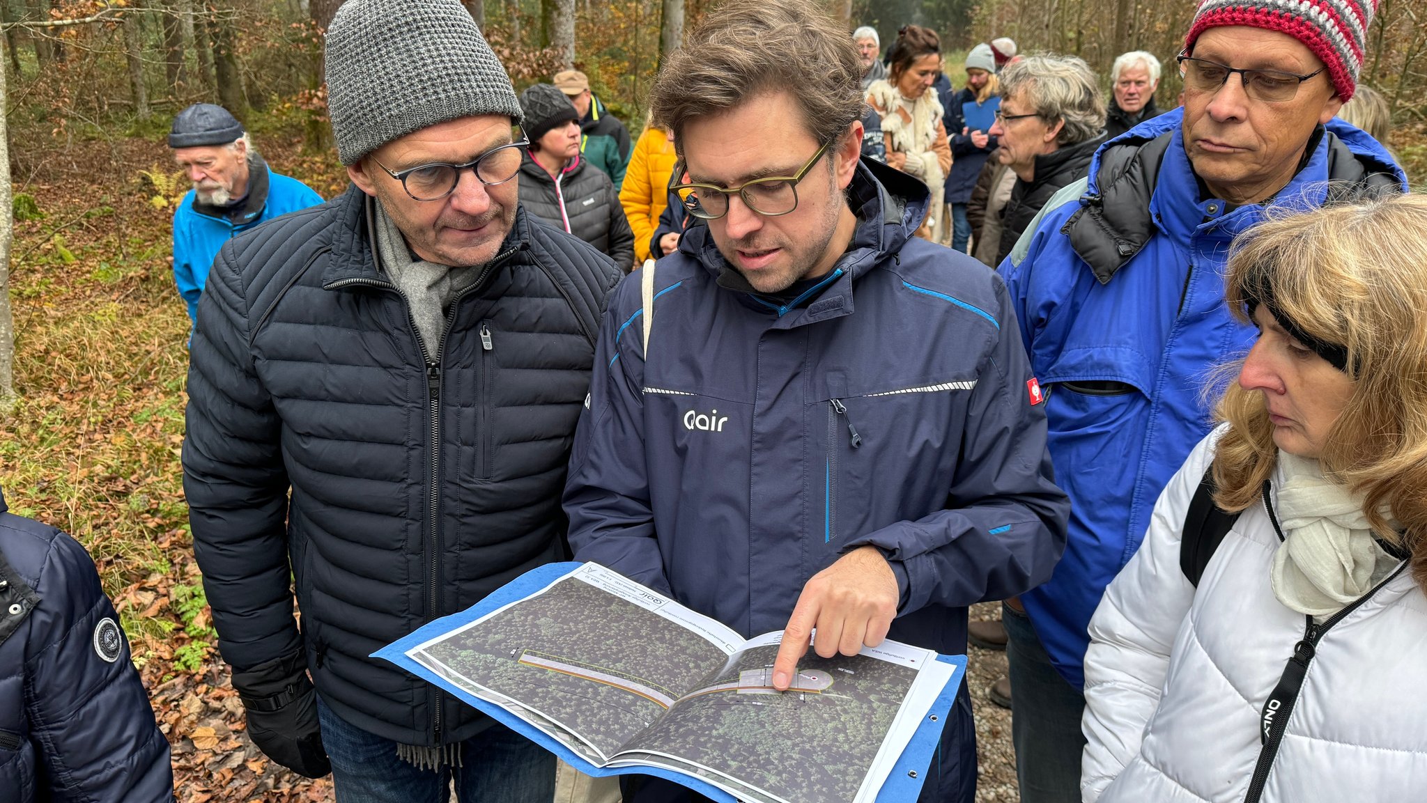 Ein Mann mit einer Jacke mit Aufschrift "Qair" hat einen Ordner in der Hand und zeigt eine Grafik. Ihm schauen Menschen über die Schulter. Im Hintergrund sind noch mehr Menschen und Wald zu sehen. 