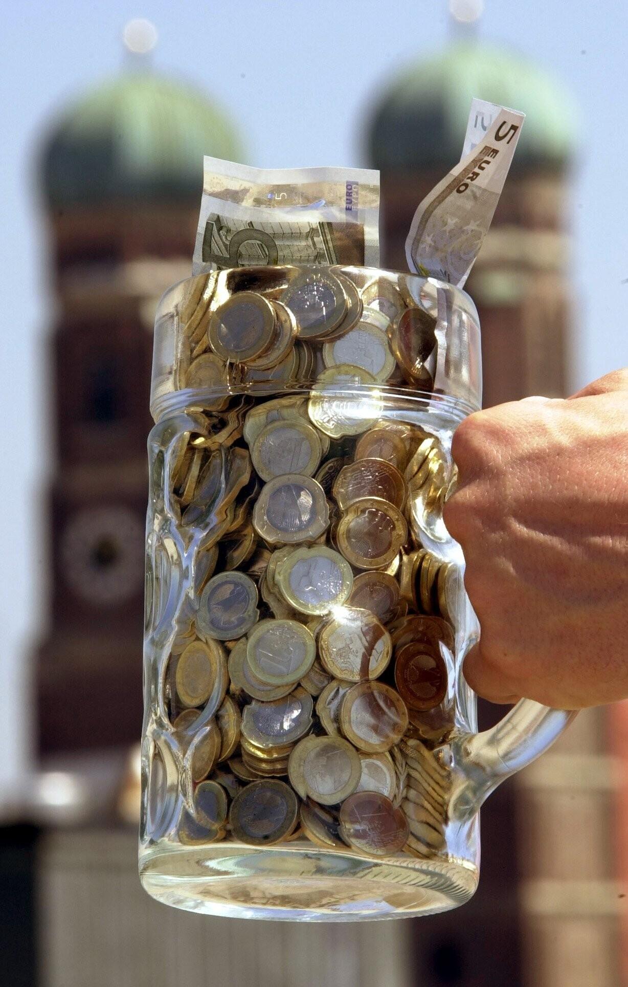 Ein Maßkrug voller Geld, dahinter die Münchner Frauenkirche.