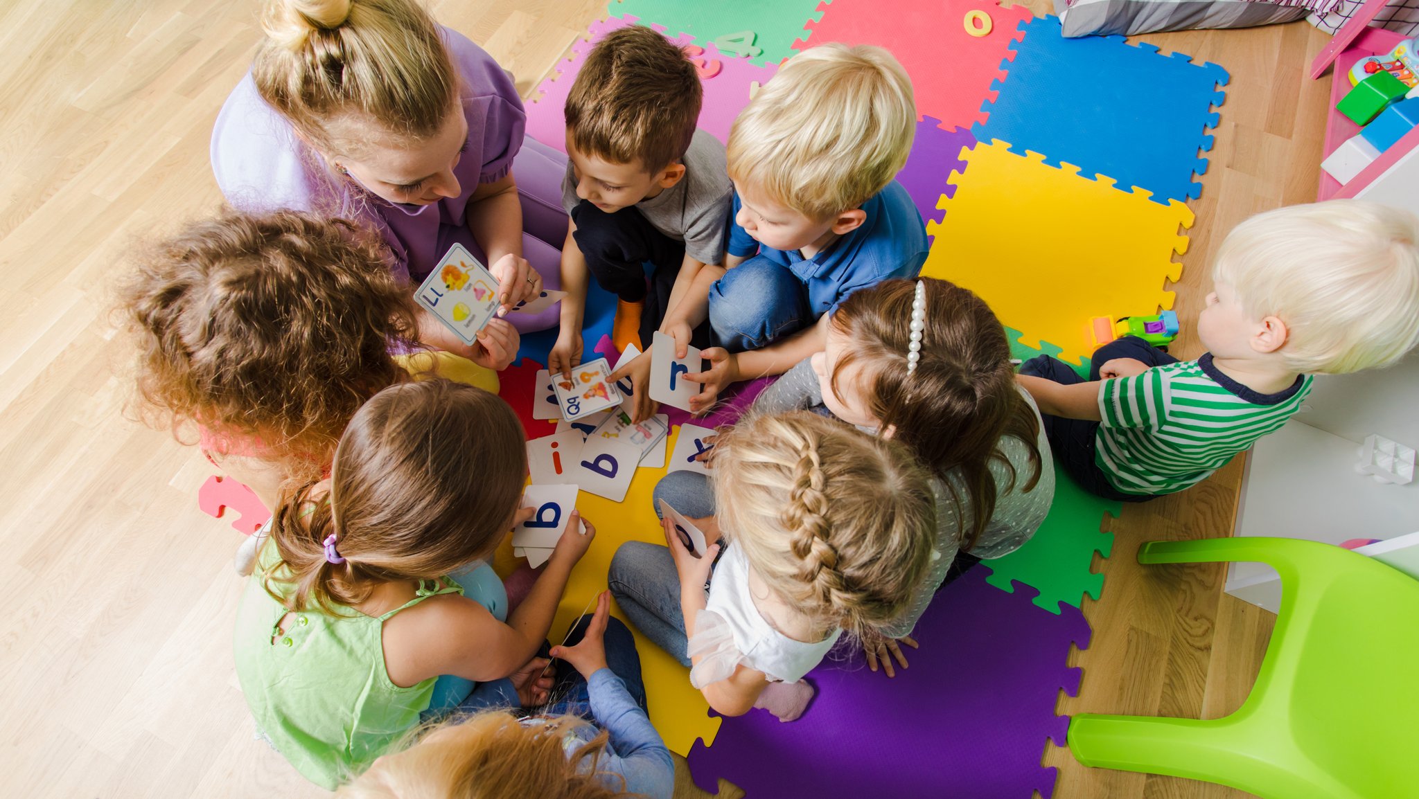 Eine Kindergartengruppe sitzt zusammen mit der Erzieherin auf dem Boden und lernt die Buchstaben.