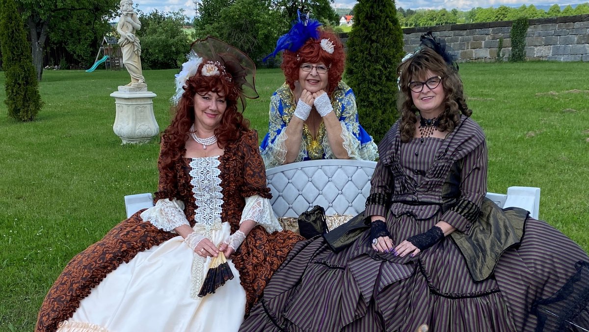 Drei Frauen sitzen in historisch anmutenden Kleidern auf einer Parkbank