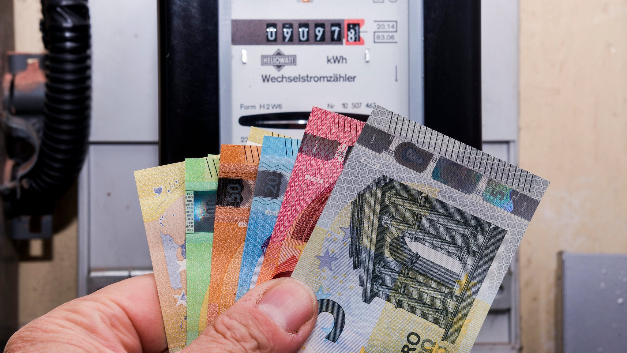 Symbolbild eines Wechselstromzählers, davor eine Hand, die je einen Fünf-, Zehn-, 20-, 50-, 100- und 200-Euro-Geldschein aufgefächert hält. Der Strompreis an der Börse sinkt, trotzdem erhalten viele Verbraucher eine Strompreiserhöhung. | Bild: picture alliance / Schoening | Schoening Symbolbild eines Wechselstromzählers, davor eine Hand, die je einen Fünf-, Zehn-, 20-, 50-, 100- und 200-Euro-Geldschein aufgefächert hält. Der Strompreis an der Börse sinkt, trotzdem erhalten viele Verbraucher eine Strompreiserhöhung.