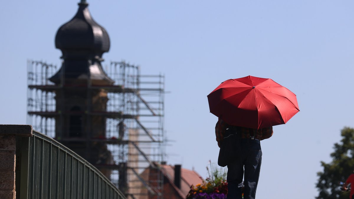 ARCHIV (19.07.2022): Ein Mann geht mit einem roten Sonnenschirm über die Alte Mainbrücke. Seit 2015 hält Kitzingen den bayerischen Temperaturrekord.