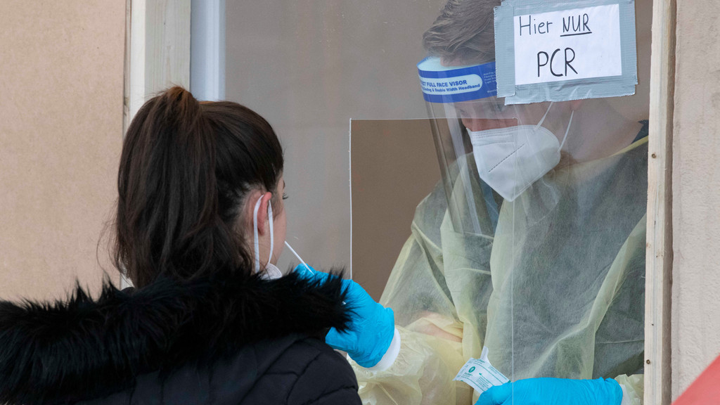 Eine junge Frau steht mit heruntergezogener FFP2-Maske an einem Fenster einer Corona-Teststation, um einen PCR-Test zu machen. Hinter dem Fenster steht ein Mann in Schutzkleidung und macht mit einem Stäbchen einen Abstrich.