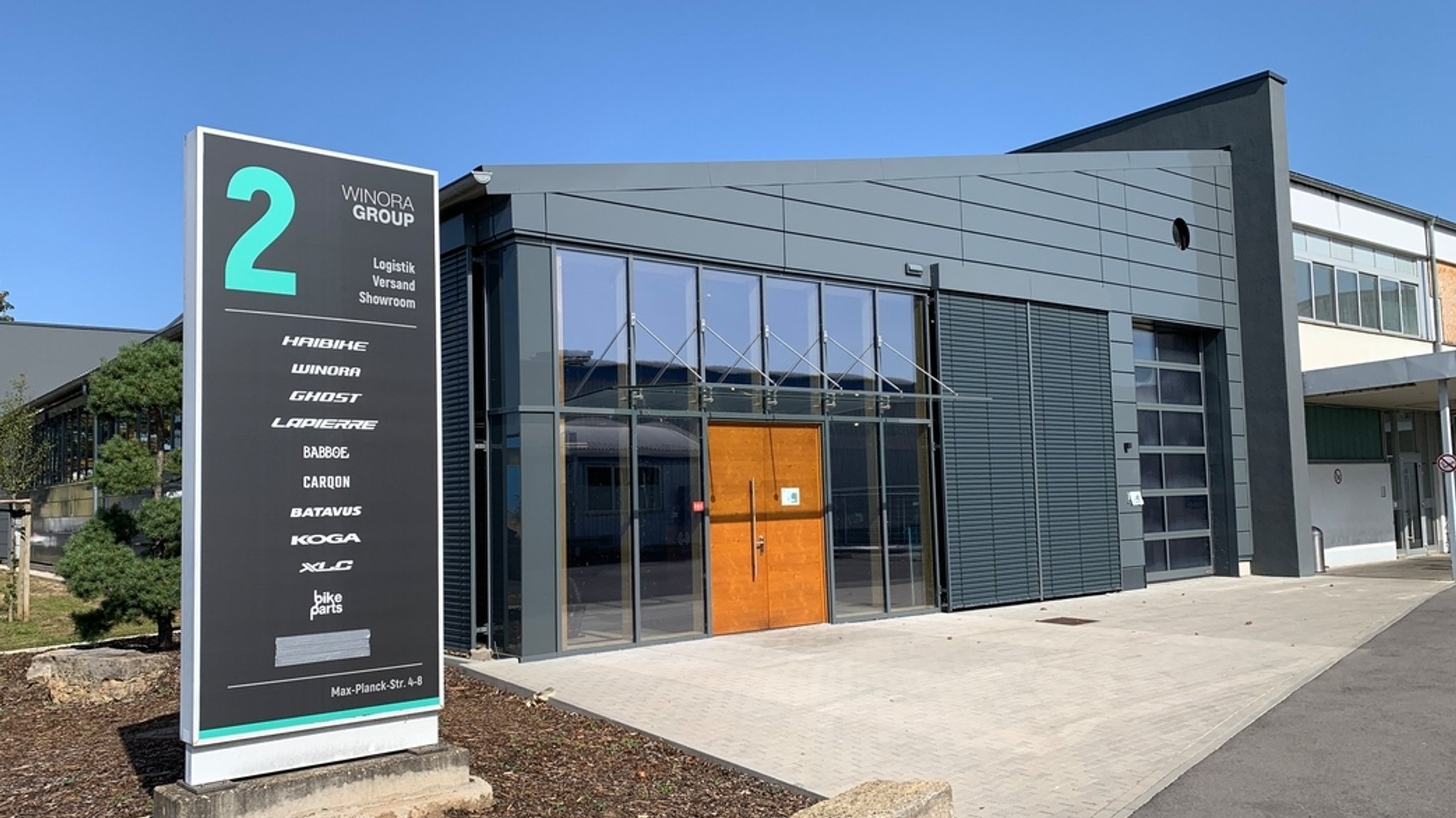 Zu sehen ist ein Gebäude der Winora Group in Sennfeld. Im Vordergrund steht ein großes Hinweisschild mit der Aufschrift „WINORA GROUP“ sowie den Marken Haibike, Winora, Ghost und weiteren Fahrradmarken. Dahinter befindet sich ein modernes, grau verkleidetes Gebäude mit großflächigen Fenstern und einem Eingangsbereich mit Holztür. Auf dem Schild ist „Logistik, Versand, Showroom“ angegeben. 