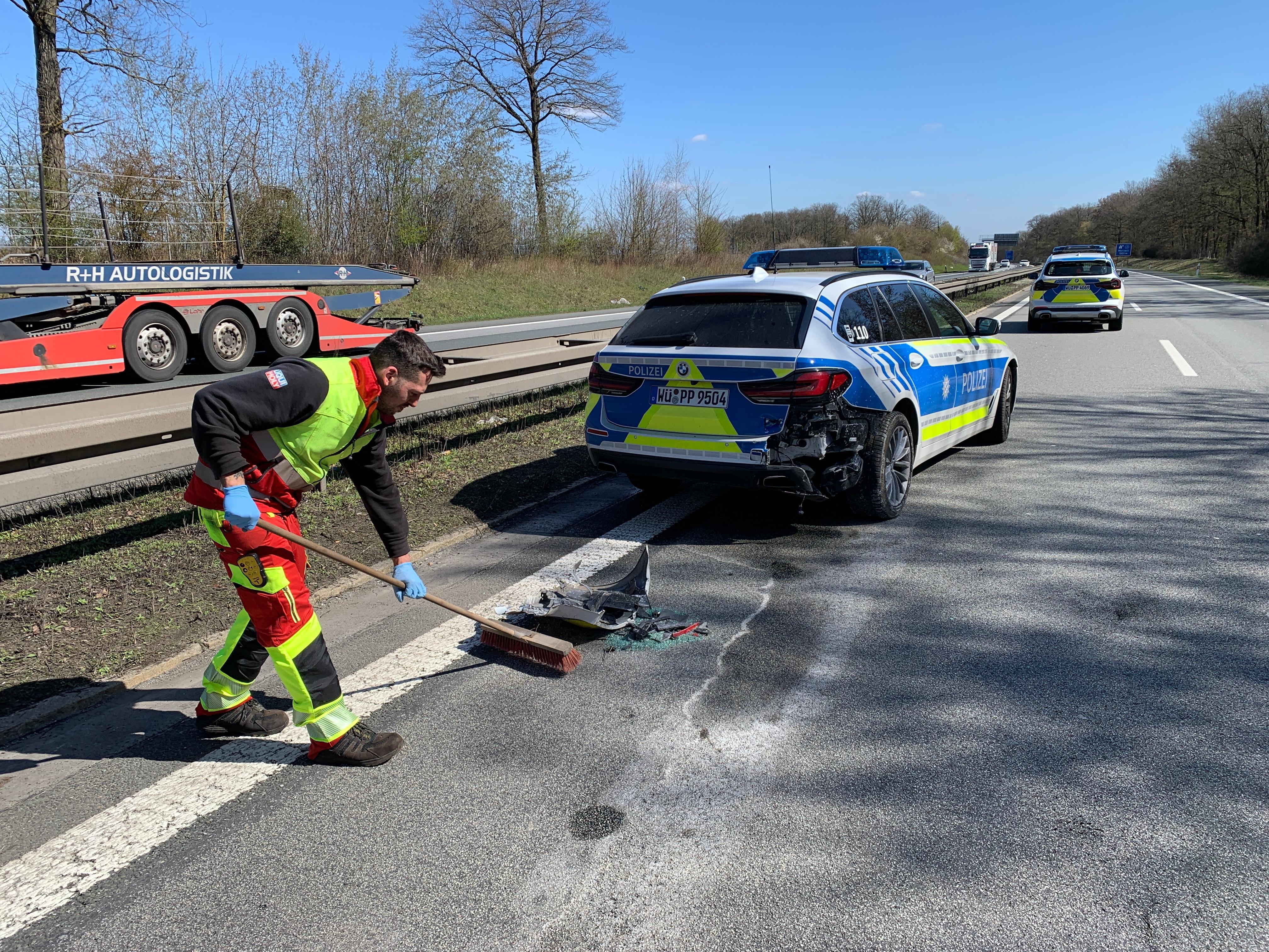 Auf einer Autobahn steht ein beschädigtes Polizeiauto. Ein Mann kehrt Teile zusammen.