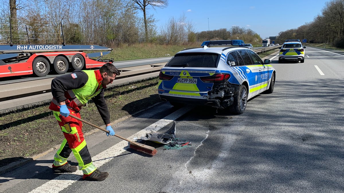 Auf einer Autobahn steht ein beschädigtes Polizeiauto. Ein Mann kehrt Teile zusammen.