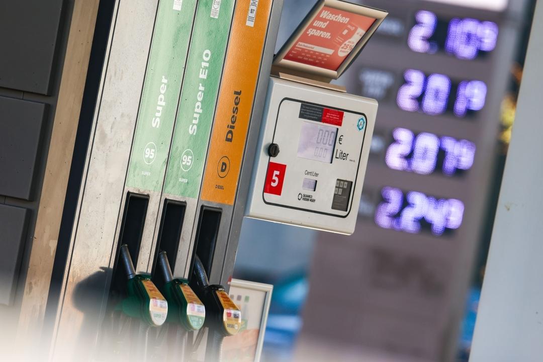 Zapfpistolen stecken in einer Zapfsäule für Super, Super E10 und Diesel, während im Hintergrund die Spritpreise auf einer Anzeige stehen.