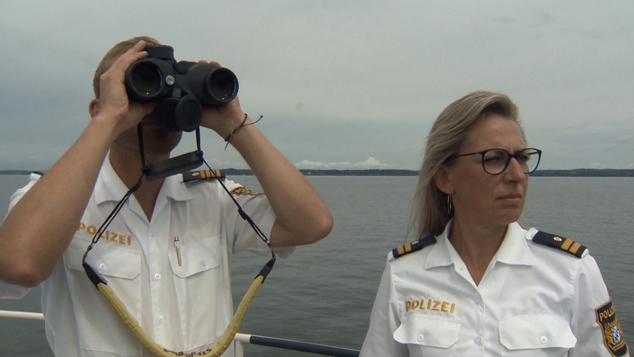 Polizist mit Fernglas und Polizistin beobachten von einem Boot aus das Wasser