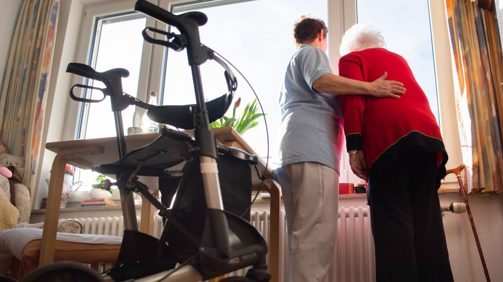 (Symbolbild) Eine Pflegerin (l) und eine Heimbewohnerin schauen zusammen aus einem Fenster im Wohnbereich des Pflegeheims. Im Vordergrund steht ein Rollator. | Bild: dpa-Bildfunk/Tom Weller (Symbolbild) Eine Pflegerin (l) und eine Heimbewohnerin schauen zusammen aus einem Fenster im Wohnbereich des Pflegeheims. Im Vordergrund steht ein Rollator.