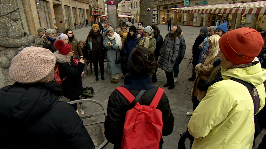 Mehrere Frauen stehen in der Fußgängerzone im Kreis um eine Frau herum, die etwas erzählt. Links von den Frauen ist eine Figur aus Stein zu sehen. 