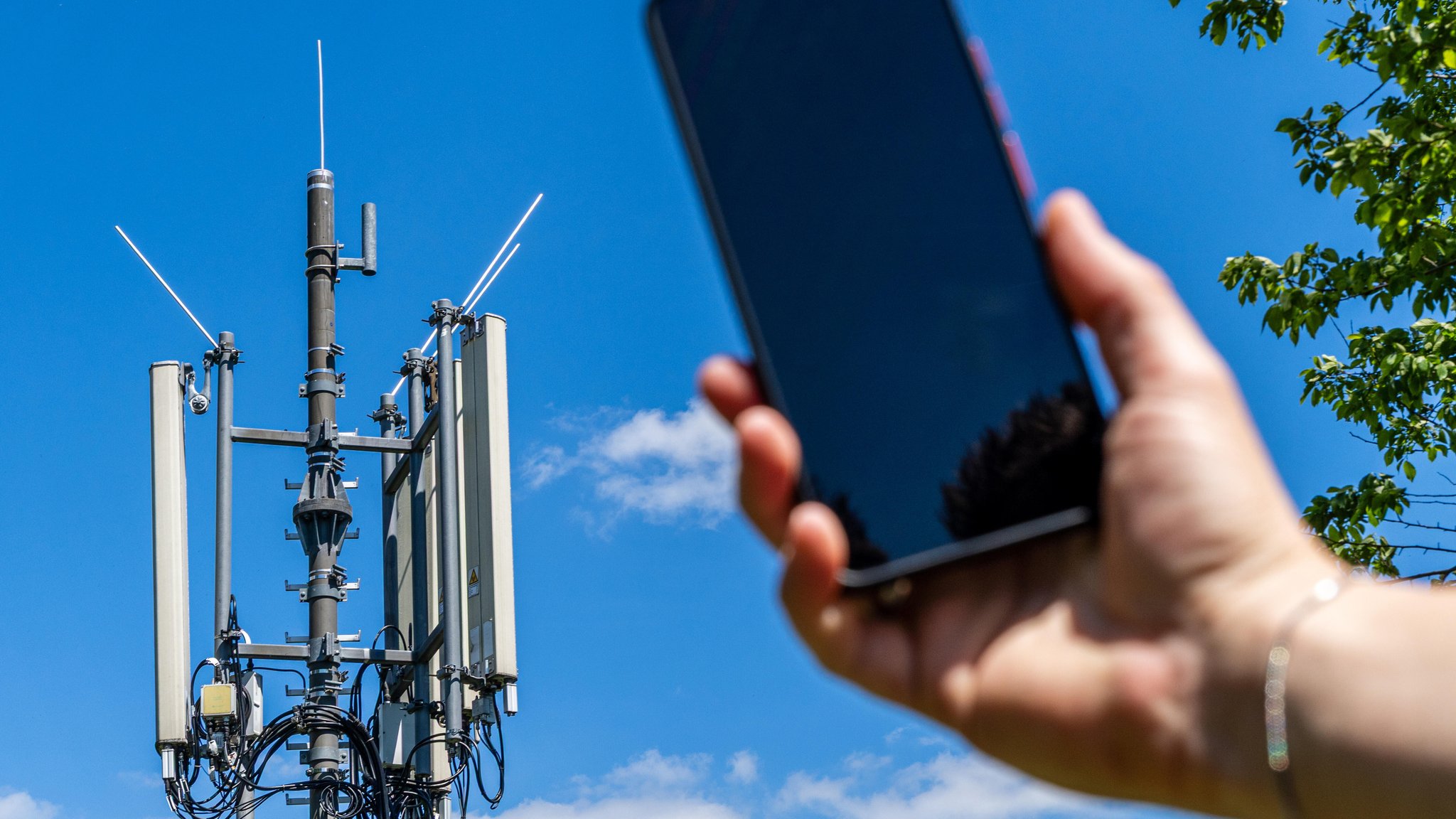 (Symbolbild) Hand hält ein Smartphone, im Hintergrund sieht man einen Handymasten 