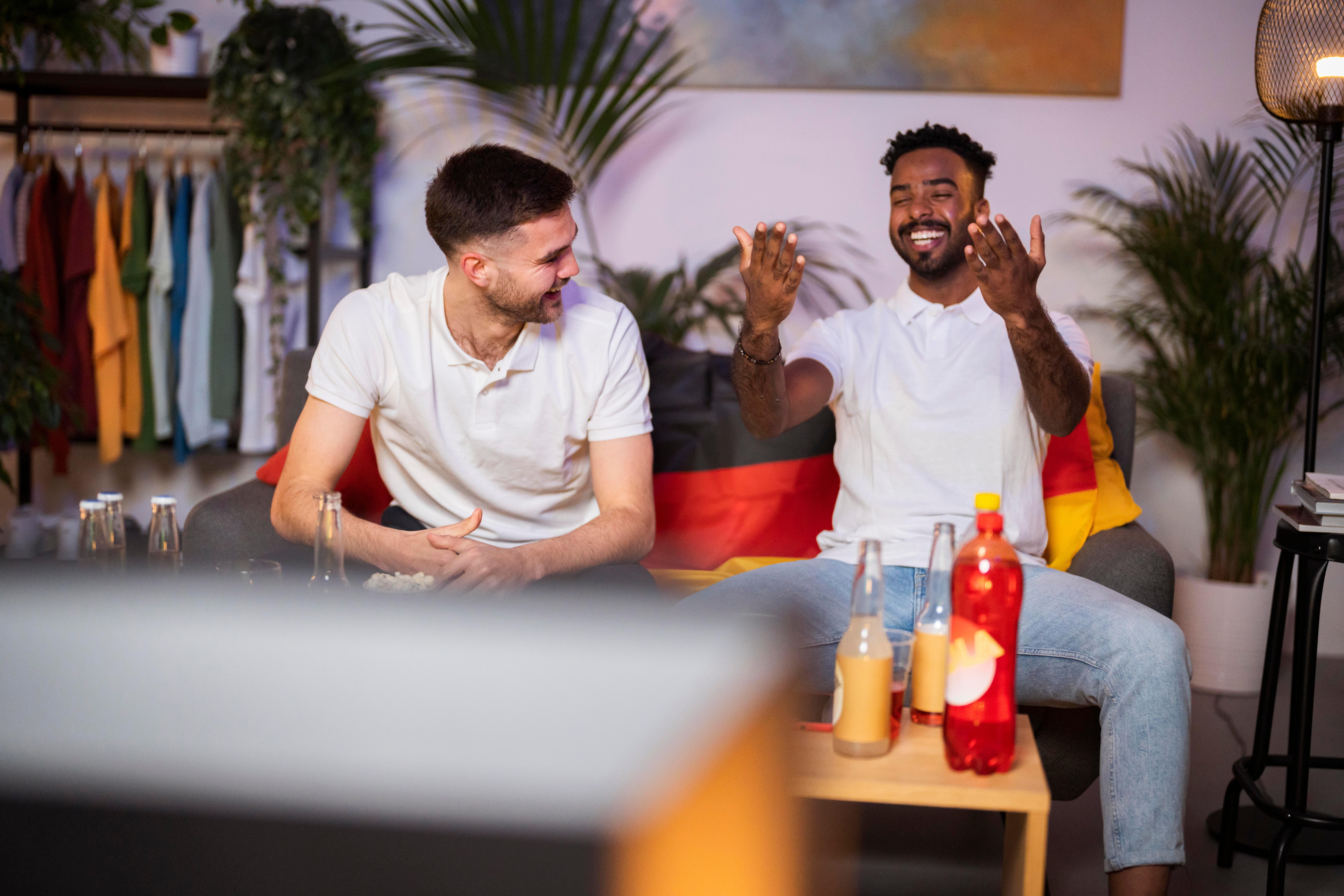 (Symbolbild) Zwei Männer sitzen auf einem Soda und schauen Fußball, hinter ihnen erkennt man eine Deutschlandflagge