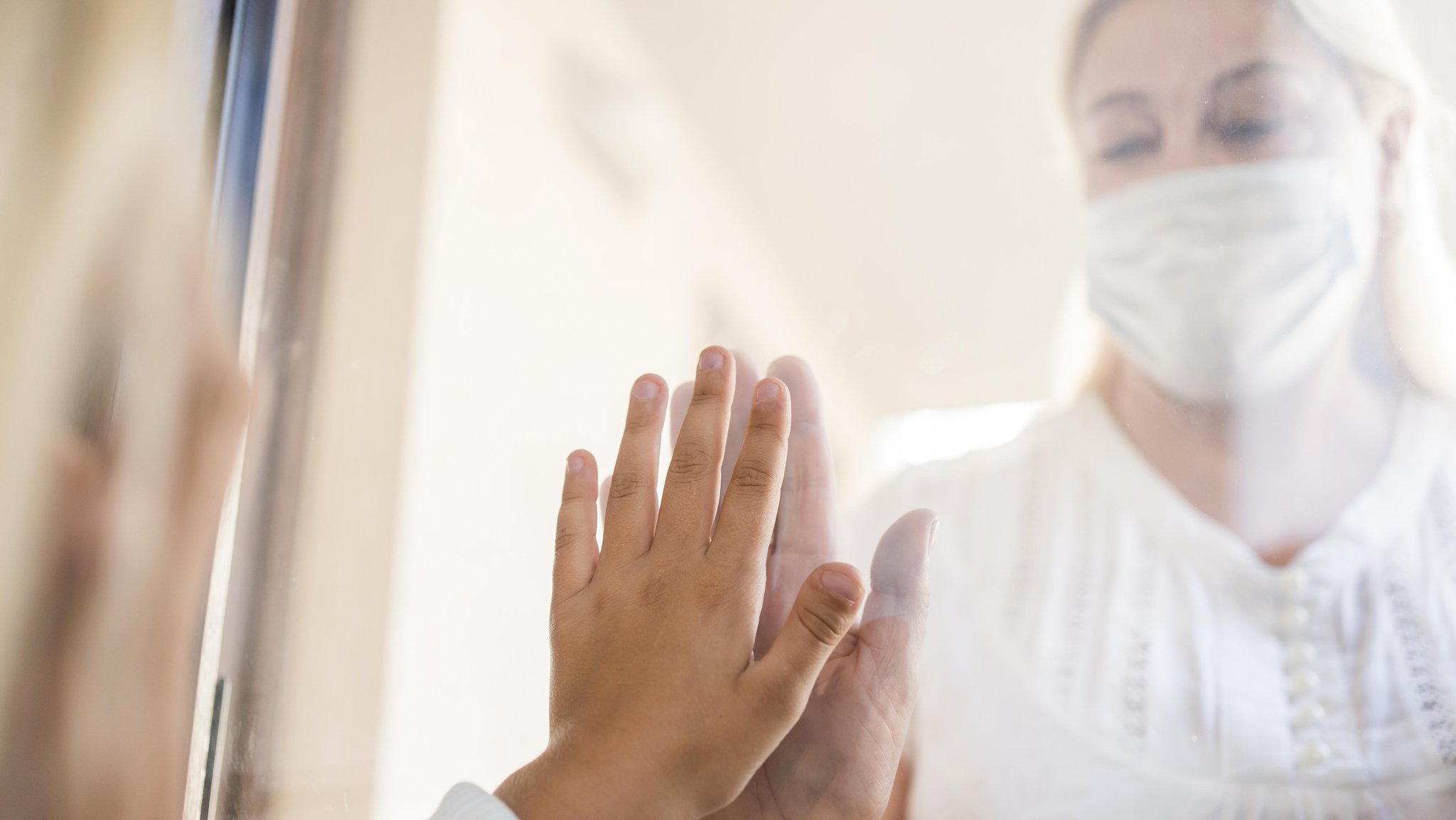 Eine Frau mit einer Maske steht an einer Glasscheibe und streckt die Hand aus (Symbolbild)