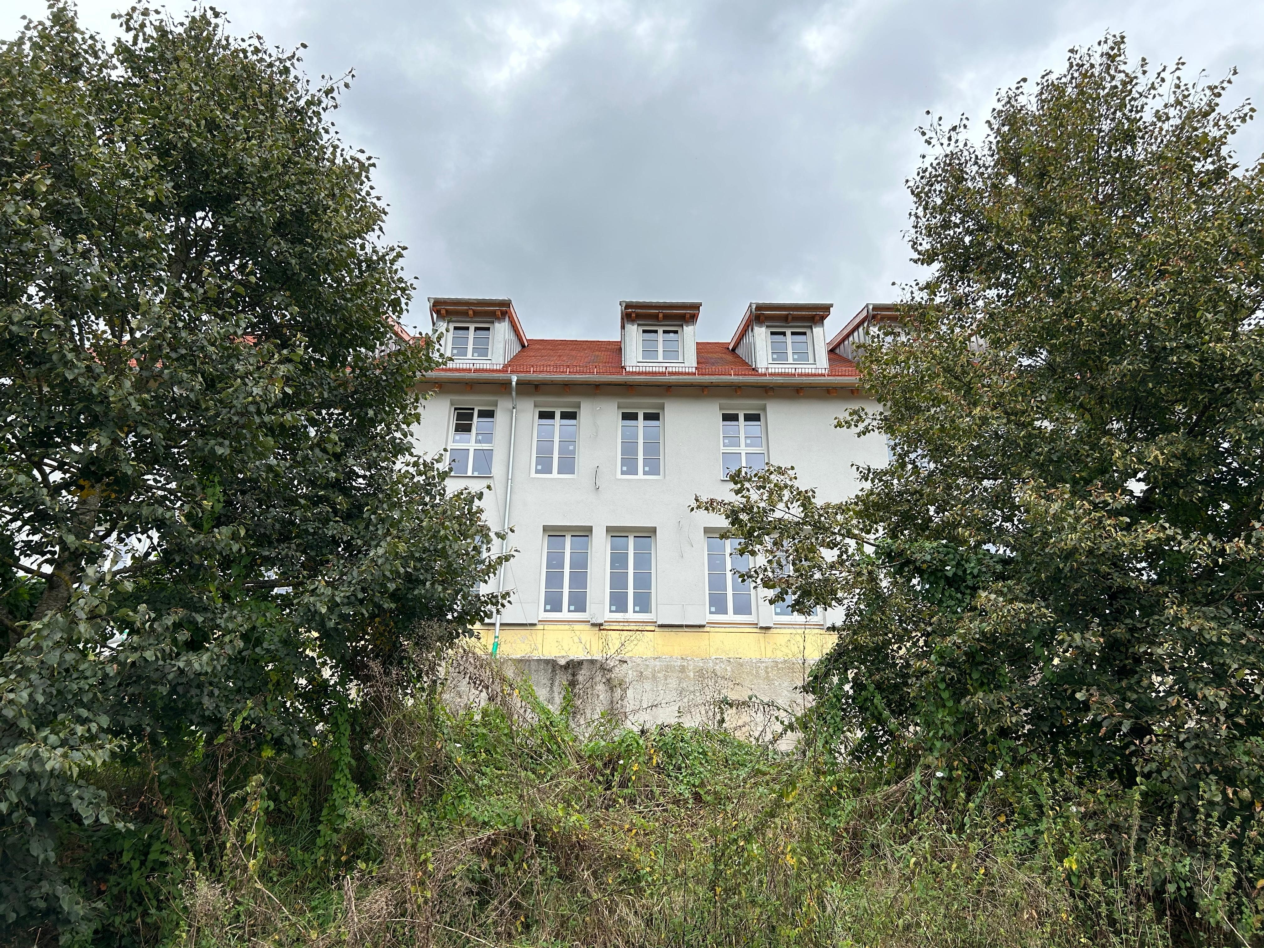 Im Vordergrund links und rechts Bäume, dazwischen im Hintergrund ein Wohnhaus