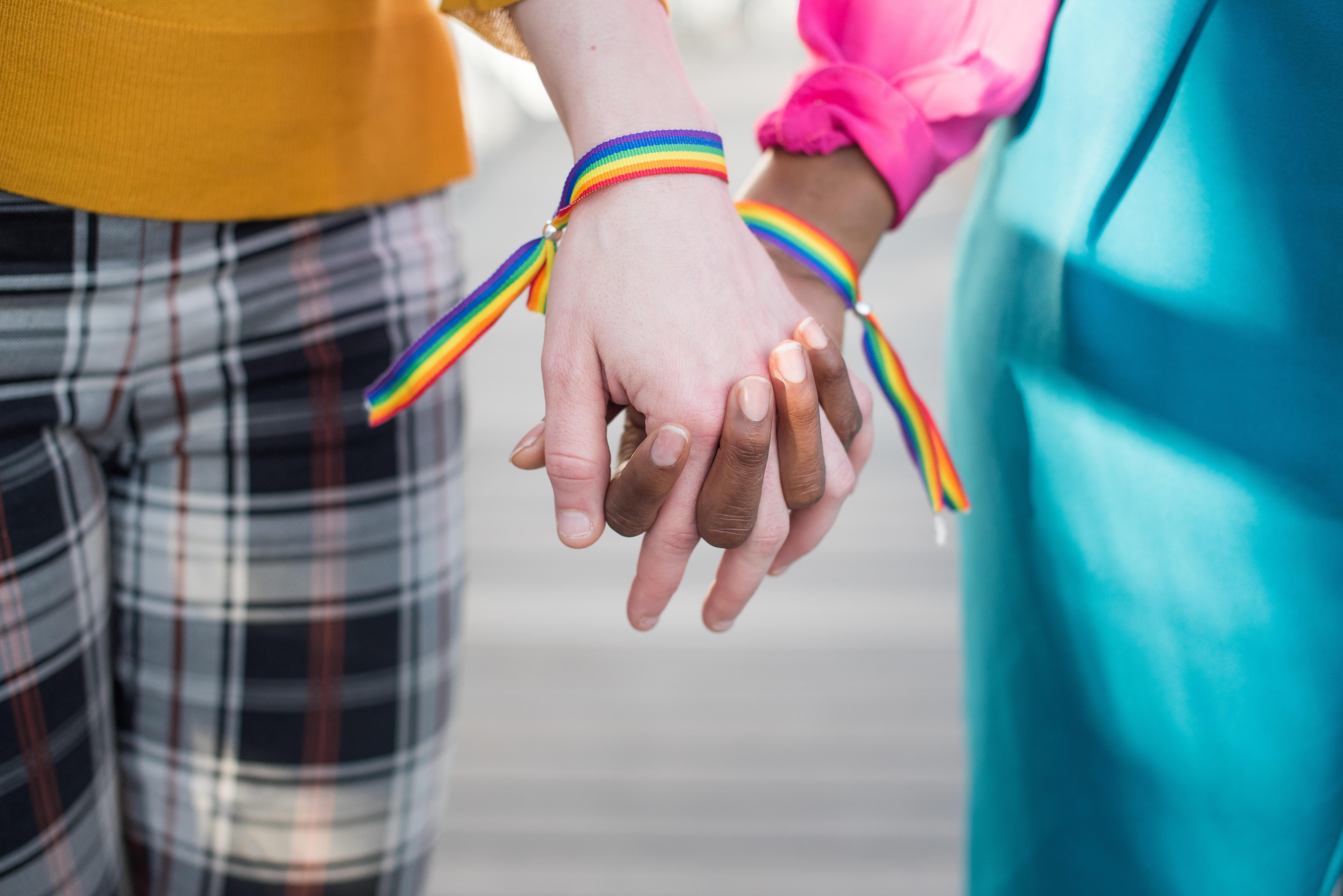 Eine weiße und eine schwarze Person stehen nebeneinander Hand in Hand, an ihren Handgelenken sind Regenbogen-Armbänder (Symbolbild)