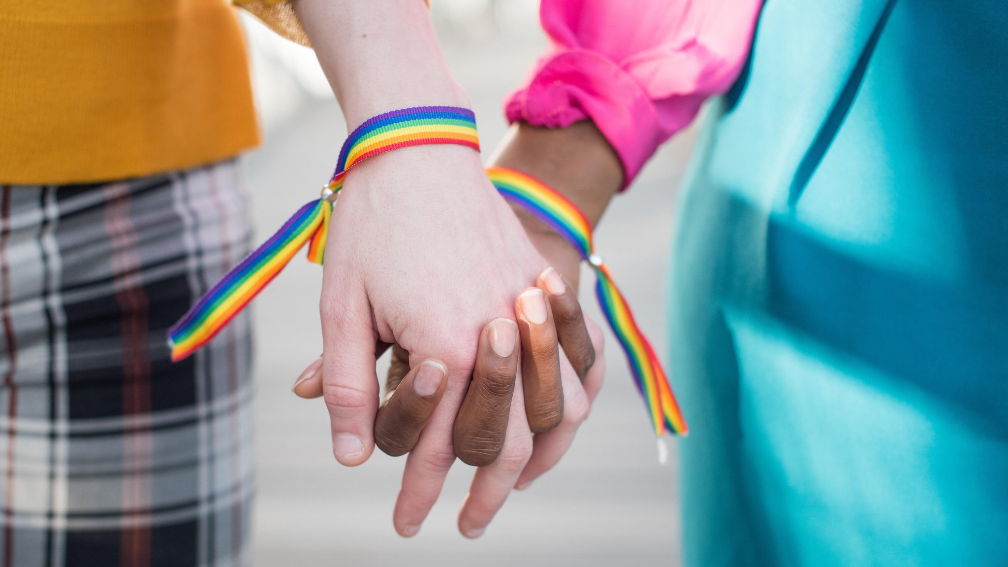 Eine weiße und eine schwarze Person stehen nebeneinander Hand in Hand, an ihren Handgelenken sind Regenbogen-Armbänder (Symbolbild)