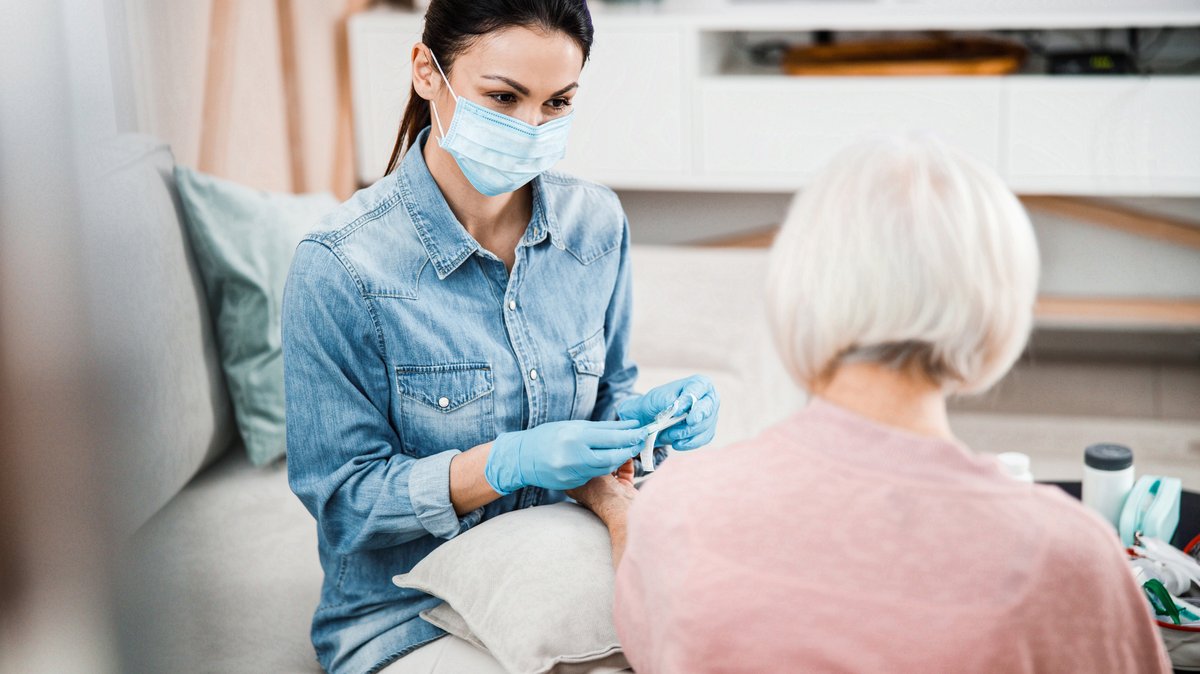 Symbolbild: Medizinisches Personal mit Mundschutz und Einweghandschuhen bei einem Hausbesuch bei einer älteren Dame.