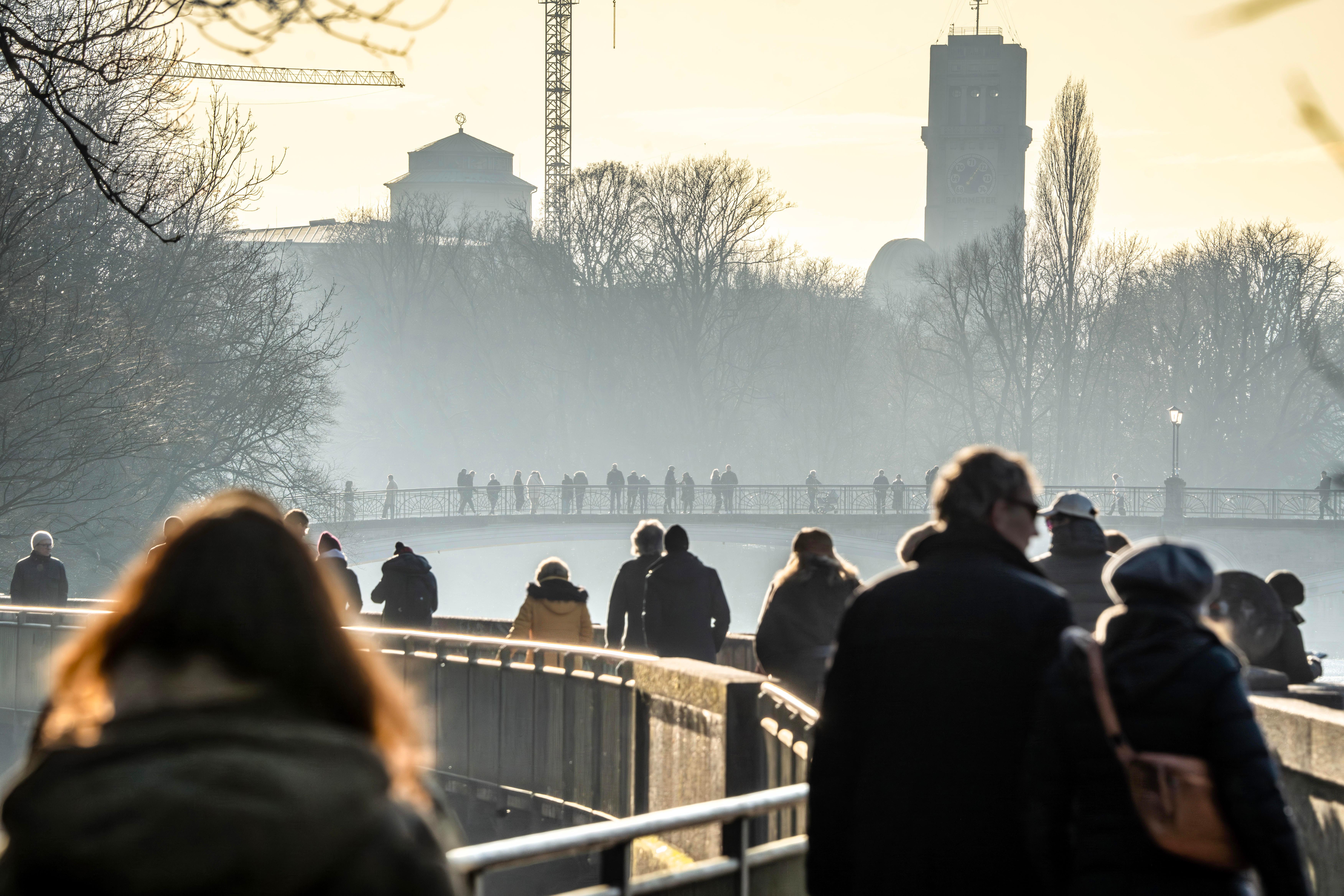 Winterlicht gekleidete Menschen laufen über eine Brücke in einem Park, im Hintergrund scheint die Sonne und es zieht Nebel vom Fluss darunter auf.