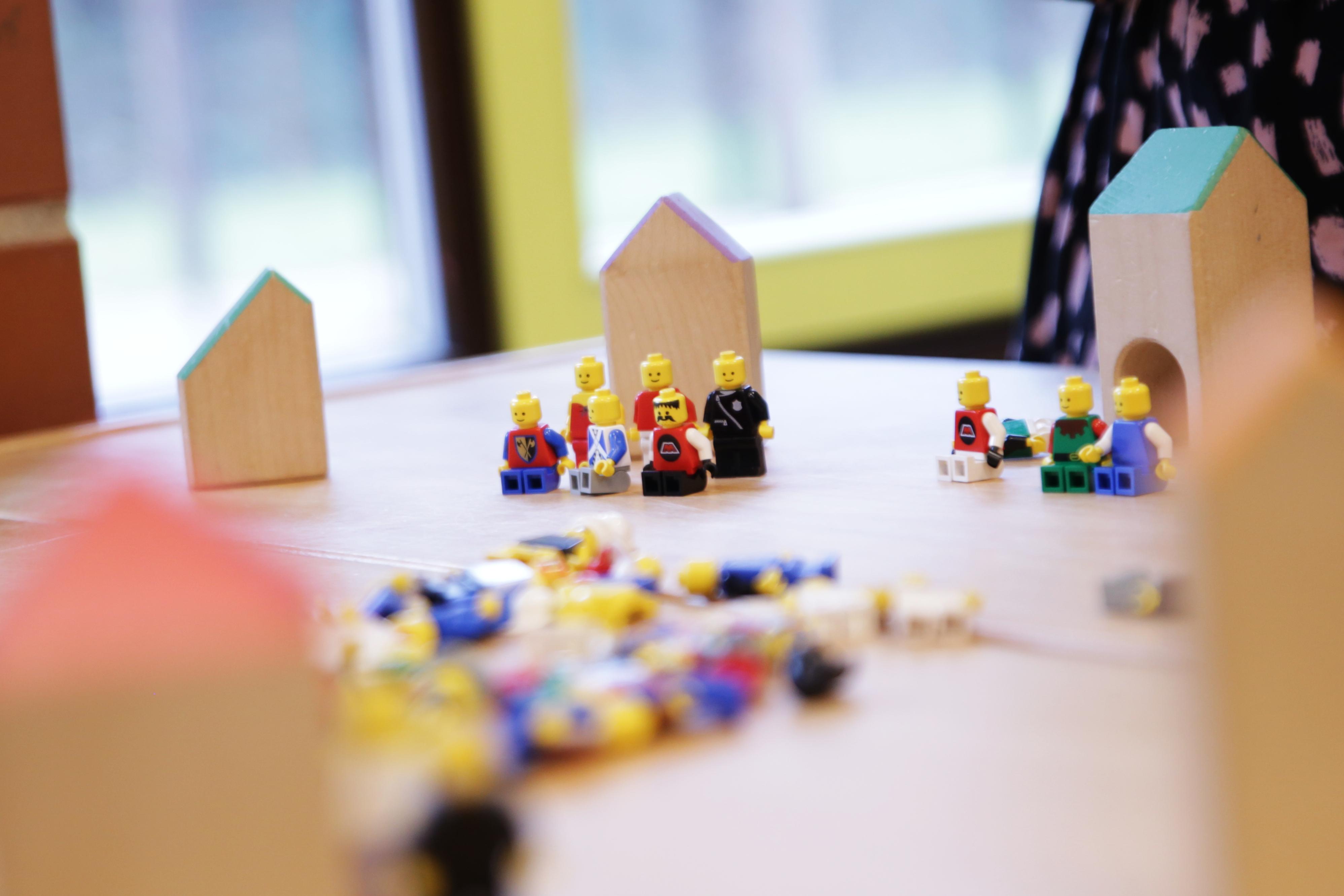 Legofiguren sitzen, stehen und liegen in einem Kindergarten auf einem Tisch. Dazu Holzklötze, die wie Häuser aussehen.