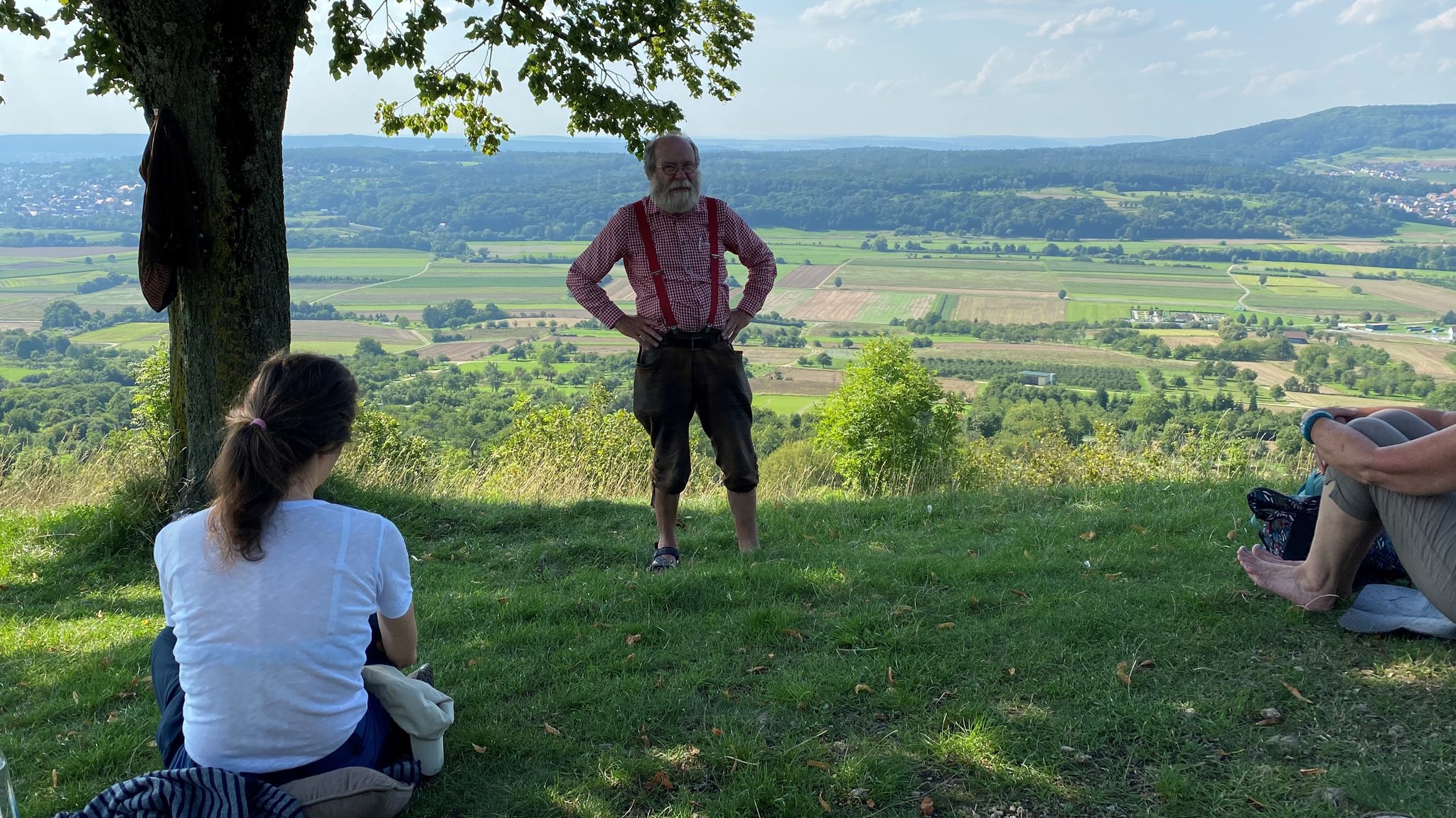 Ein Mann steht unter einem Baum und zwei Frauen sitzen im Gras vor ihm.