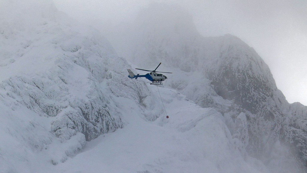 Ein Hubschrauber der Bergwacht fliegt entlang einer vereisten und verschneiten Flanke des Hochkalter. An einem Seil hängt ein tonnenförmiges Suchgerät. 