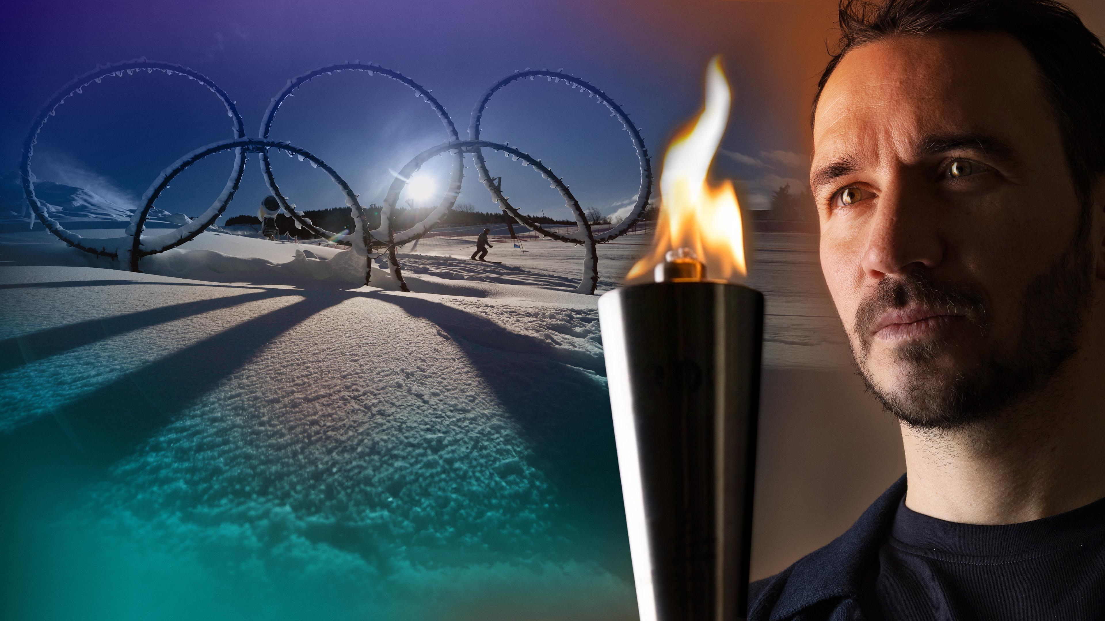 Felix Neureuther blickt mit nachdenklichem Blick auf eine olympische Fackel in seiner Hand, im Hintergrund die im Schnee stehenden Olympischen Ringe im Stelvio Ski Center in Bormio.
