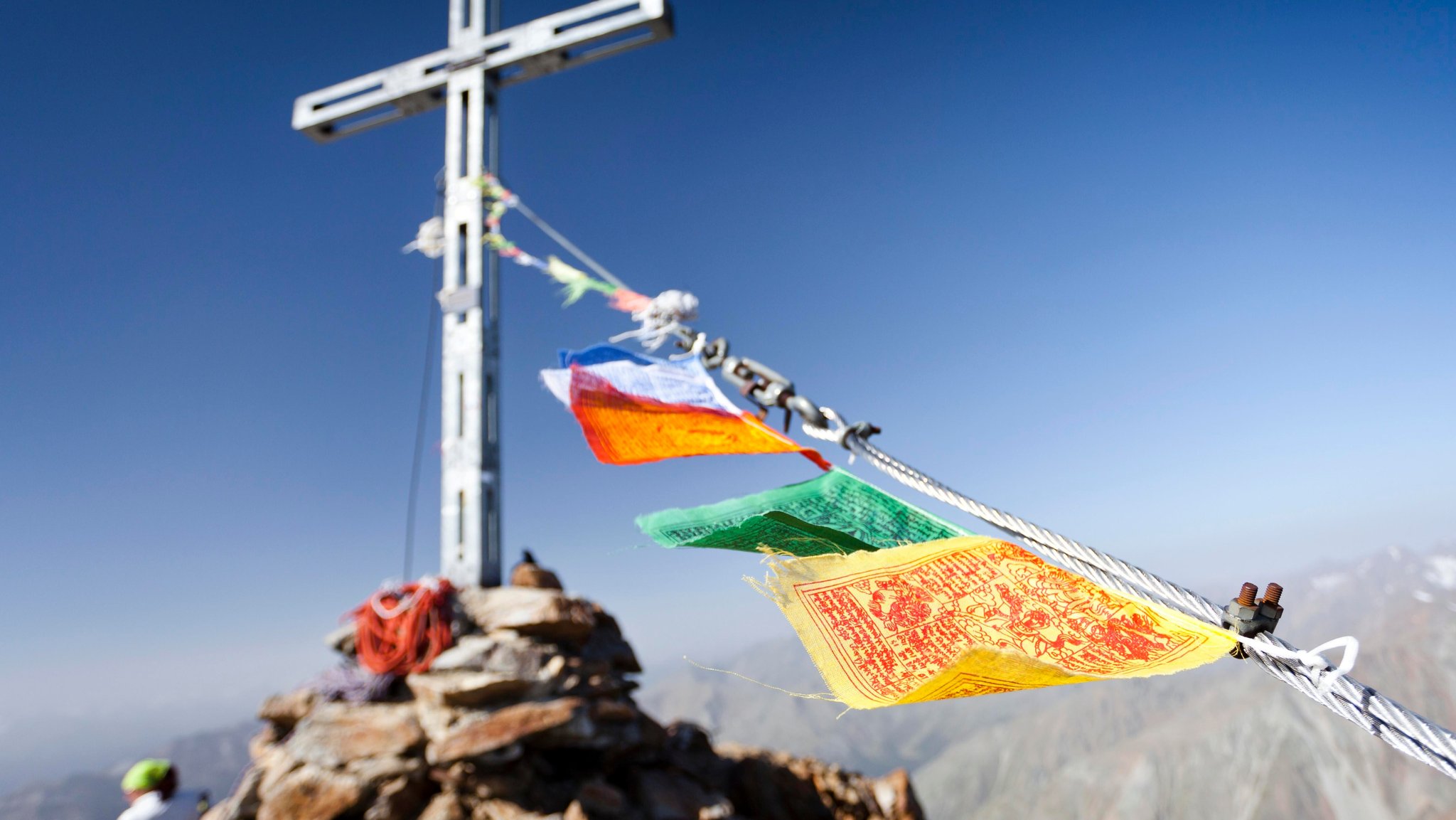 Bunte Tibet-Fähnchen wehen im Wind auf einem Berggipfel an einem Gipfelkreuz.