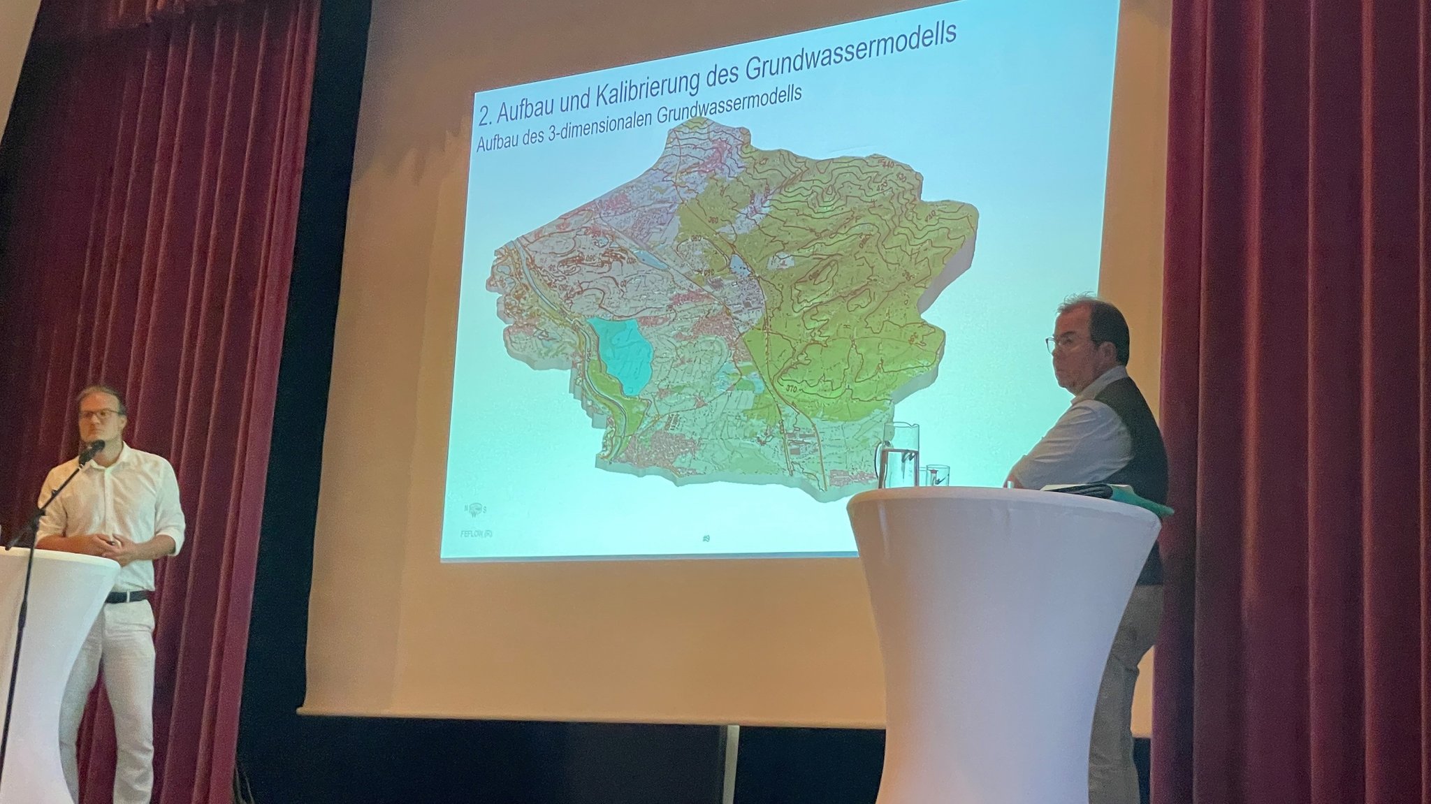 Die Bühne im Bürgersaal von Münchsmünster; links und rechts an Stehtischen zwei Experten. Zwischen ihnen hängt eine Leinwand und darauf eine Projektion eines Grundwassermodells.