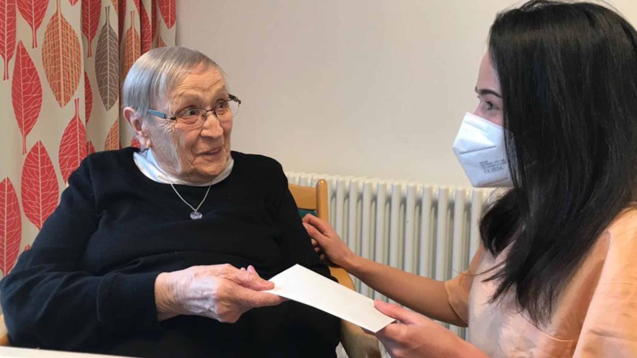 Eine junge Frau mit Maske überreicht einer Seniorin einen Brief.