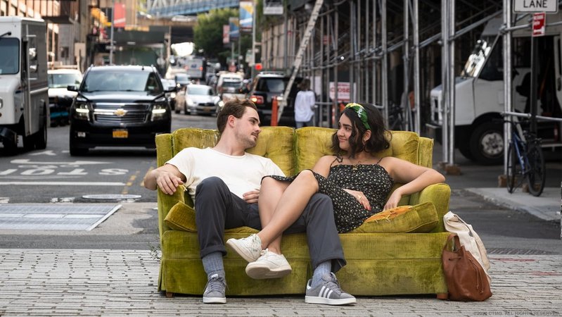 Ein junger Mann und eine junge Frau sitzen auf einem grüngelben Zweiersofa mitten auf einer befahrenen Straße: Szene aus "The Broken Hearts Gallery" (Filmszene).