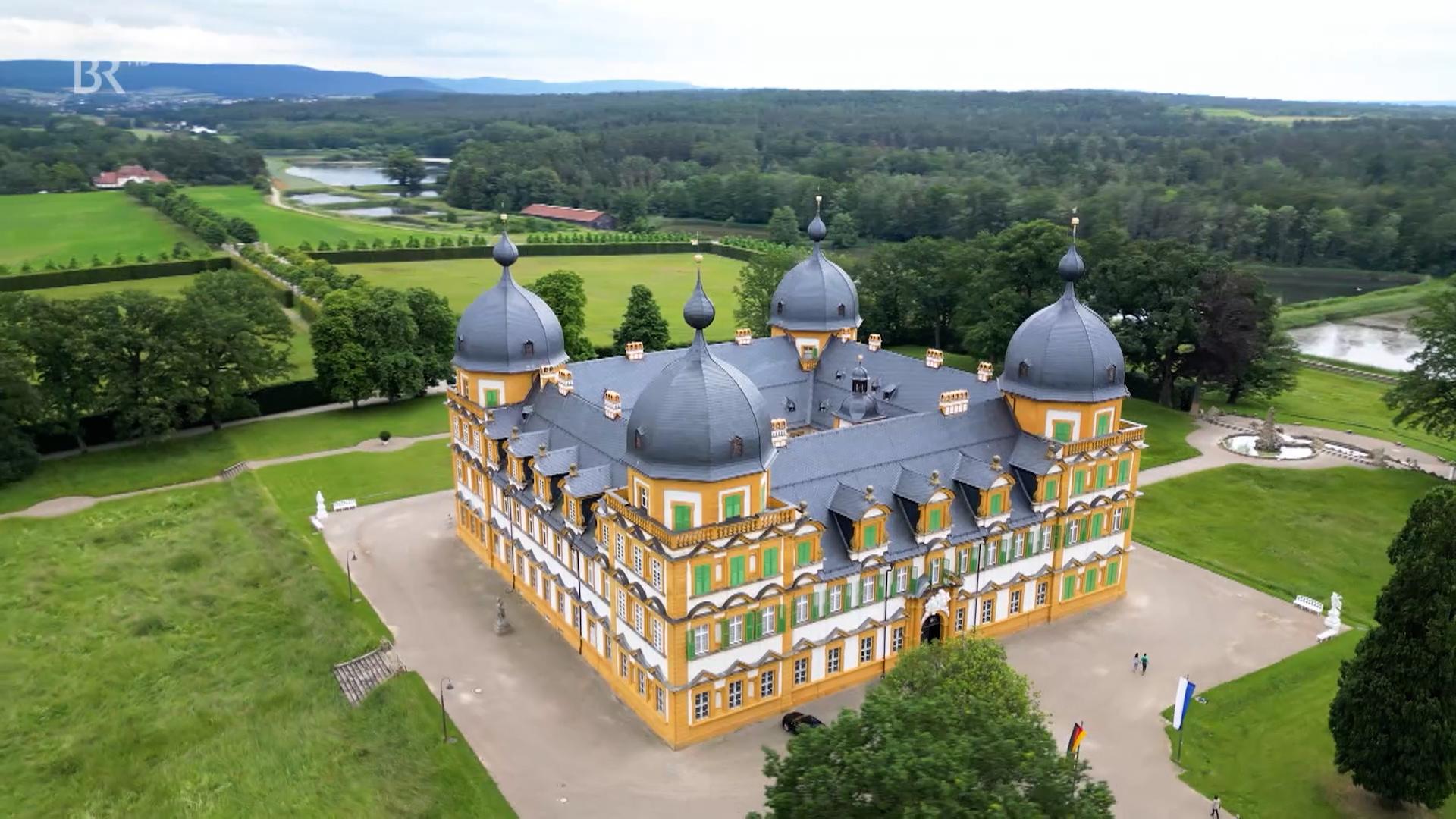 Ein Schloss Seehof mit seinen vier Türmen aus der Vogelperspektive betrachtet. Im Hintergrund Teiche und  Wald. 
