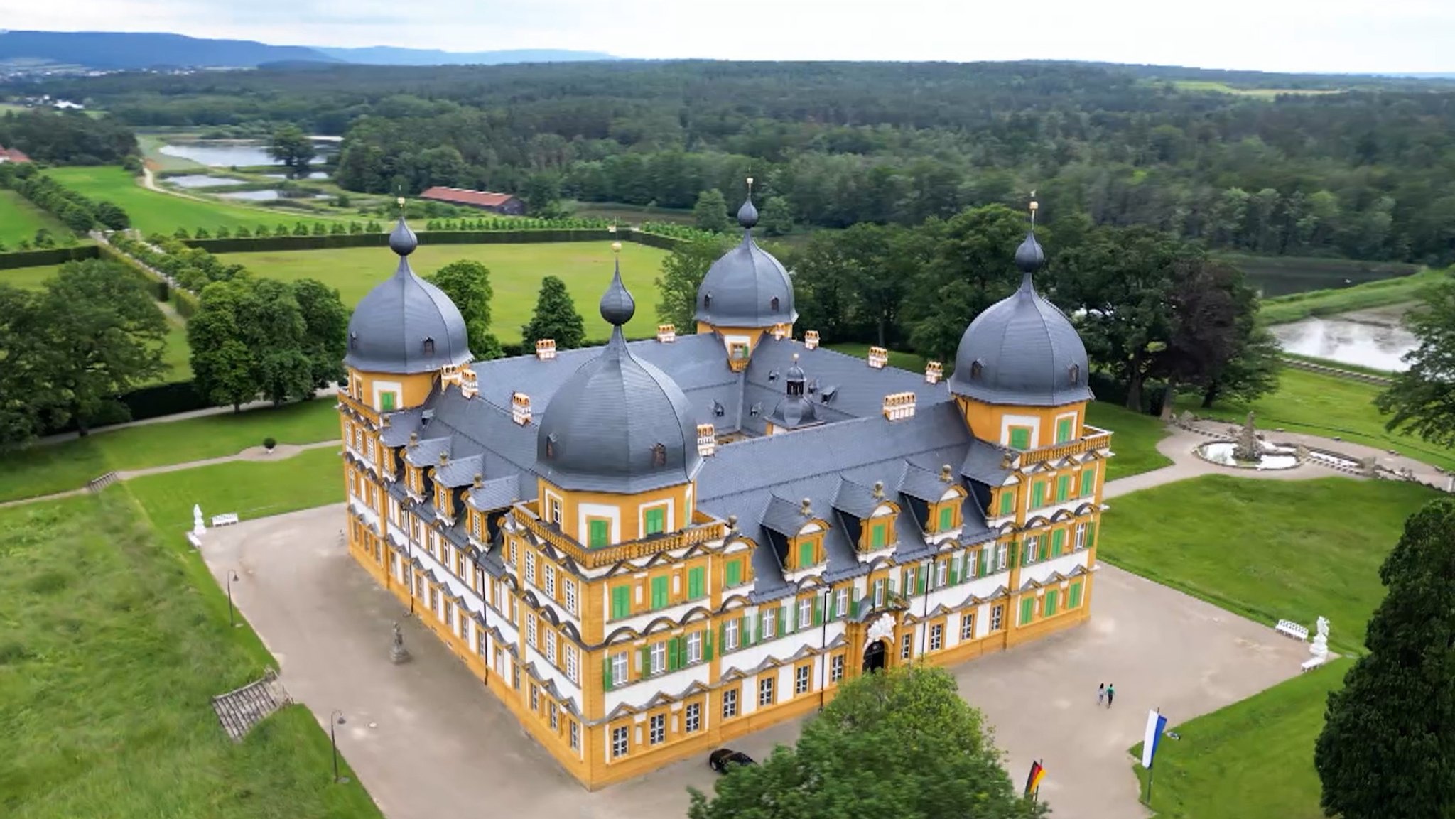 Ein Schloss Seehof mit seinen vier Türmen aus der Vogelperspektive betrachtet. Im Hintergrund Teiche und  Wald. 