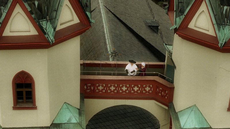 Ein Mann und eine Frau stehen in luftiger Höhe auf dem Verbindungsbau zwischen zwei Kirchtürmen.  
