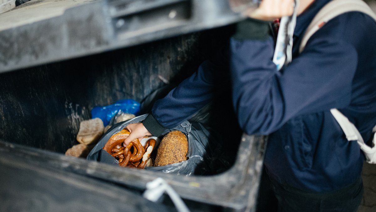 Eine Frau fischt Lebensmittel aus einer Mülltonne eines Supermarkts.