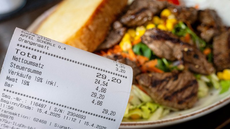 Ein Kassenzettel mit dem Mehrwertsteuersatz von 19 Prozent wird vor einen Teller mit Essen gehalten. | Bild: dpa-Bildfunk/Sina Schuldt Ein Kassenzettel mit dem Mehrwertsteuersatz von 19 Prozent wird vor einen Teller mit Essen gehalten.