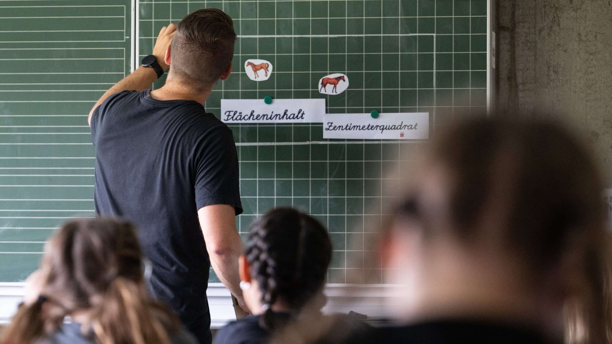 PRODUKTION - 27.06.2024, Baden-Württemberg, Neckartailfingen: Ein Lehrer macht im Unterricht einer vierten Klasse einer Grundschule einen Tafelanschrieb, während an der Tafel Zettel mit der Aufschrift «Flächeninhalt» und «Zentimeterquadrat» hängen. (zu dpa: «Hamburg vermeldet Rekord bei neu eingestellten Lehrkräften») Foto: Marijan Murat/dpa +++ dpa-Bildfunk +++