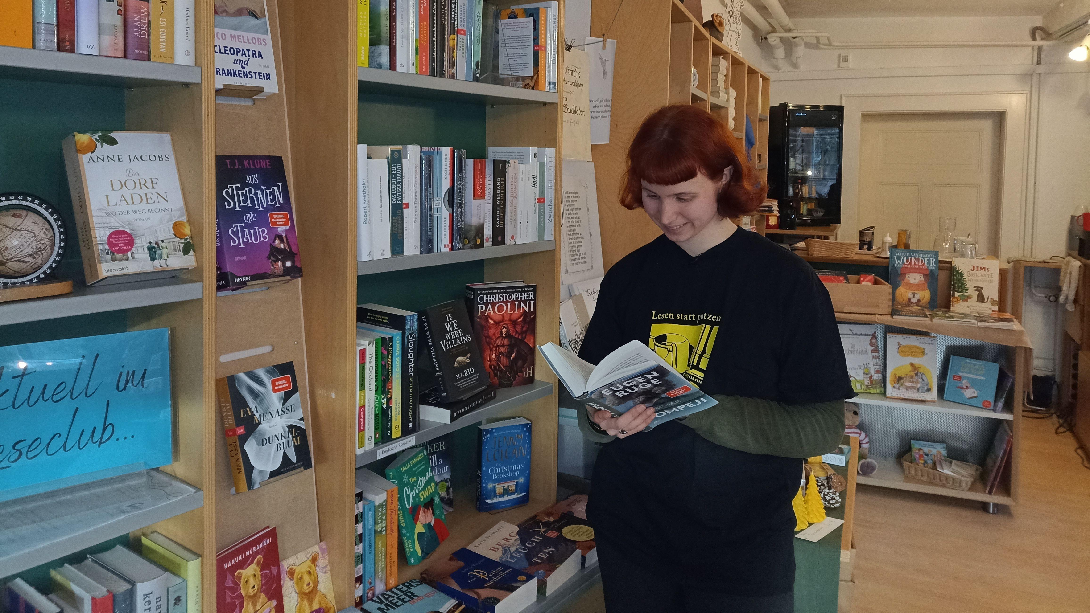 Junge Frau steht an einem Bücherregal und liest in einem Buch.