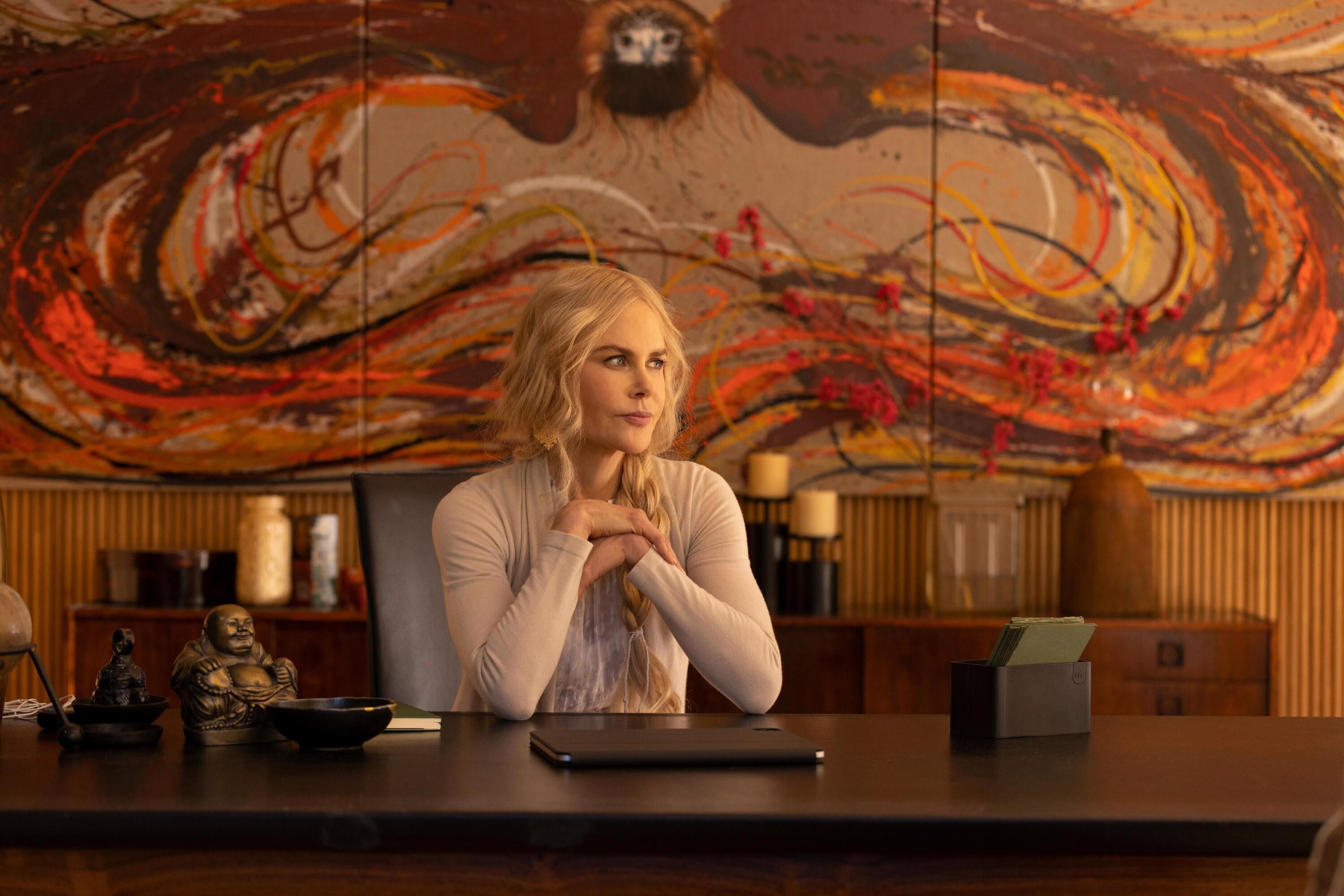Nicole Kidman als spirituelle Heilerin Masha sitzt, den Blick zur Seite gewendet, an einem großen Holztisch. Im Hintergrund ein Sideboard mit Kerzen und an der Wand ein großes schwungvolles Gemälde in Rot- und Brauntönen. Szene aus der ersten Staffel der Serie "Nine Perfect Strangers".