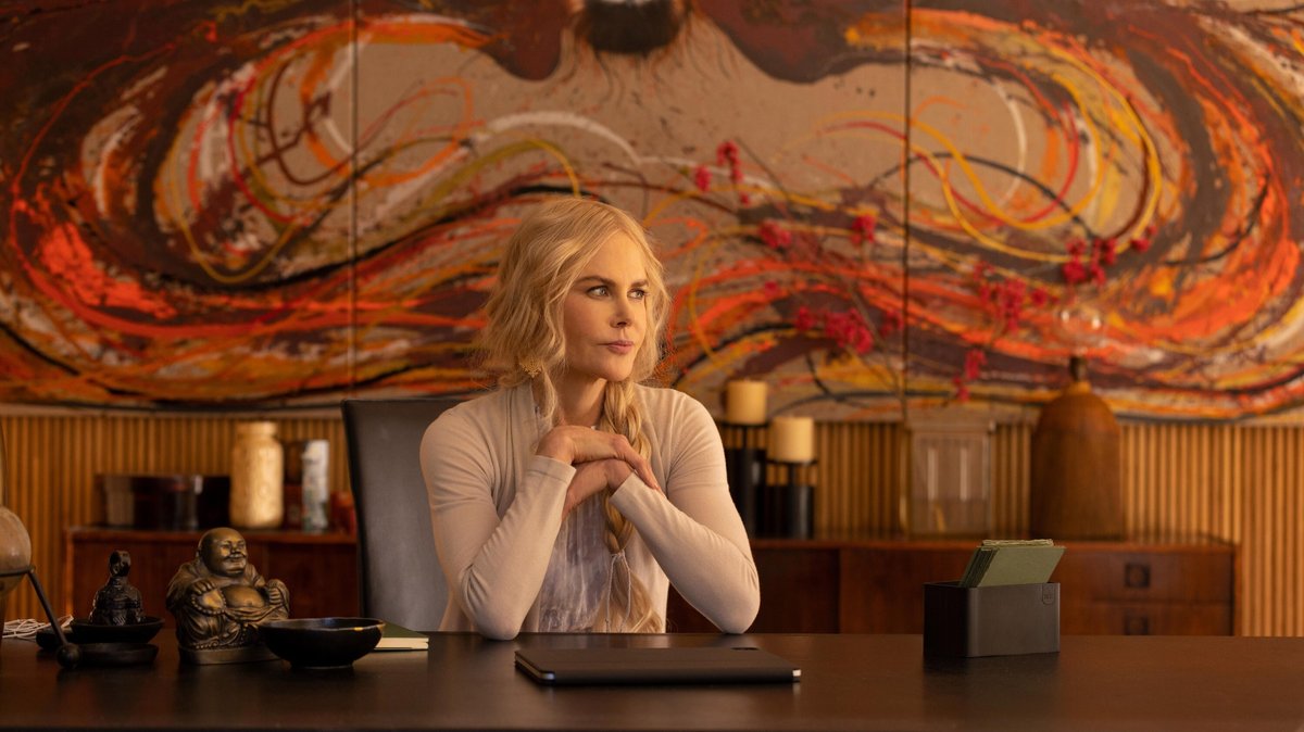 Nicole Kidman als spirituelle Heilerin Masha sitzt, den Blick zur Seite gewendet, an einem großen Holztisch. Im Hintergrund ein Sideboard mit Kerzen und an der Wand ein großes schwungvolles Gemälde in Rot- und Brauntönen. Szene aus der ersten Staffel der Serie "Nine Perfect Strangers".