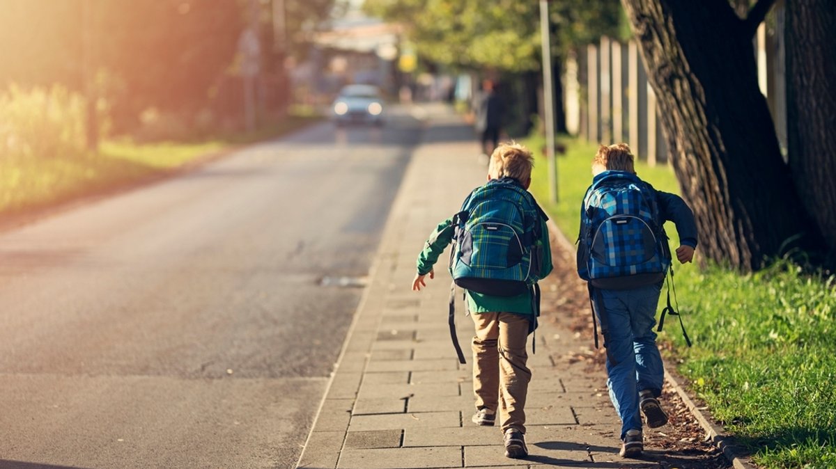 Zwei Kinder mit Schulranzen rennen eine Straße entlang. Ein Auto kommt entgegen.