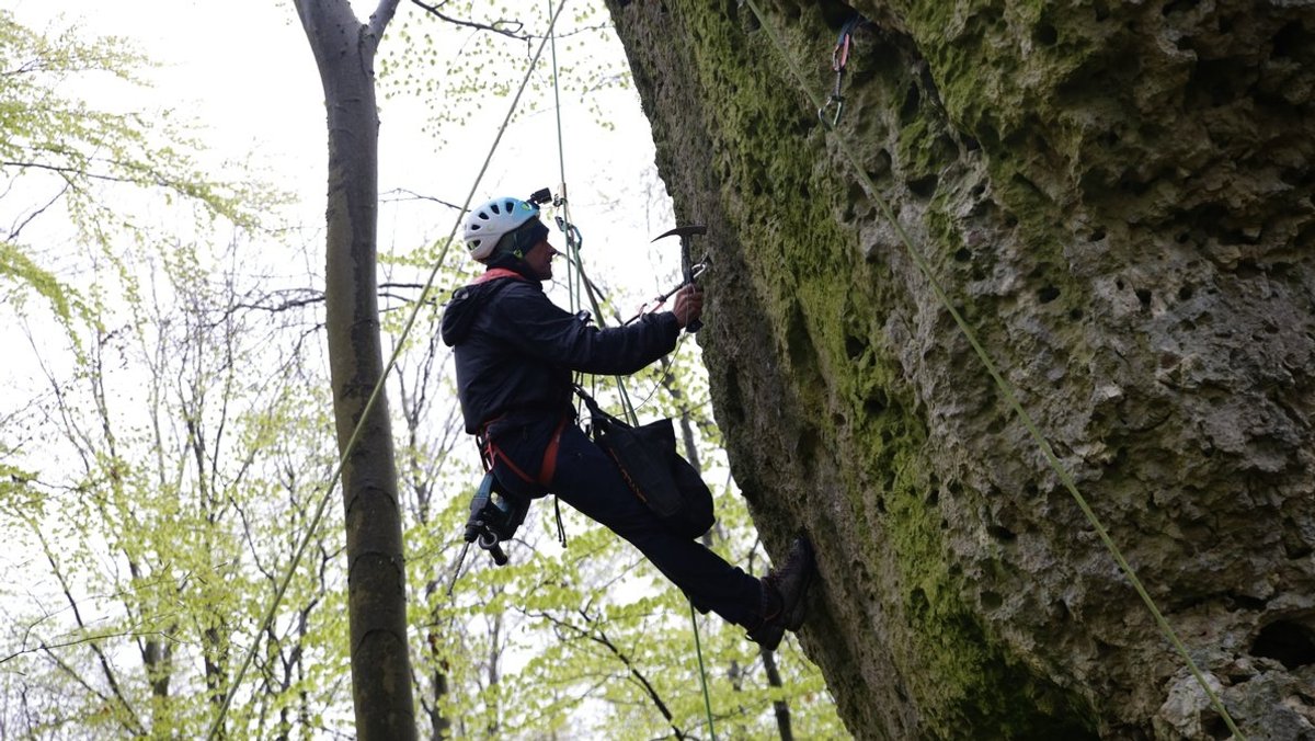 Ein Kletterer hängt an einem Seil und klopft gegen den Felsen, um seine Qualität zu prüfen. 
