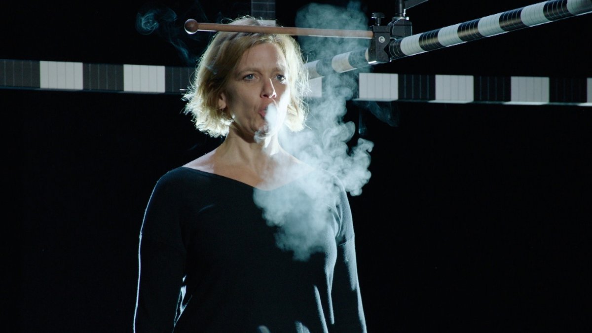 Eine Frau steht vor schwarzem Hintergrund und atmet aus. Ihr Atmen ist im Gegenlicht als Dampfwolke zu erkennen.